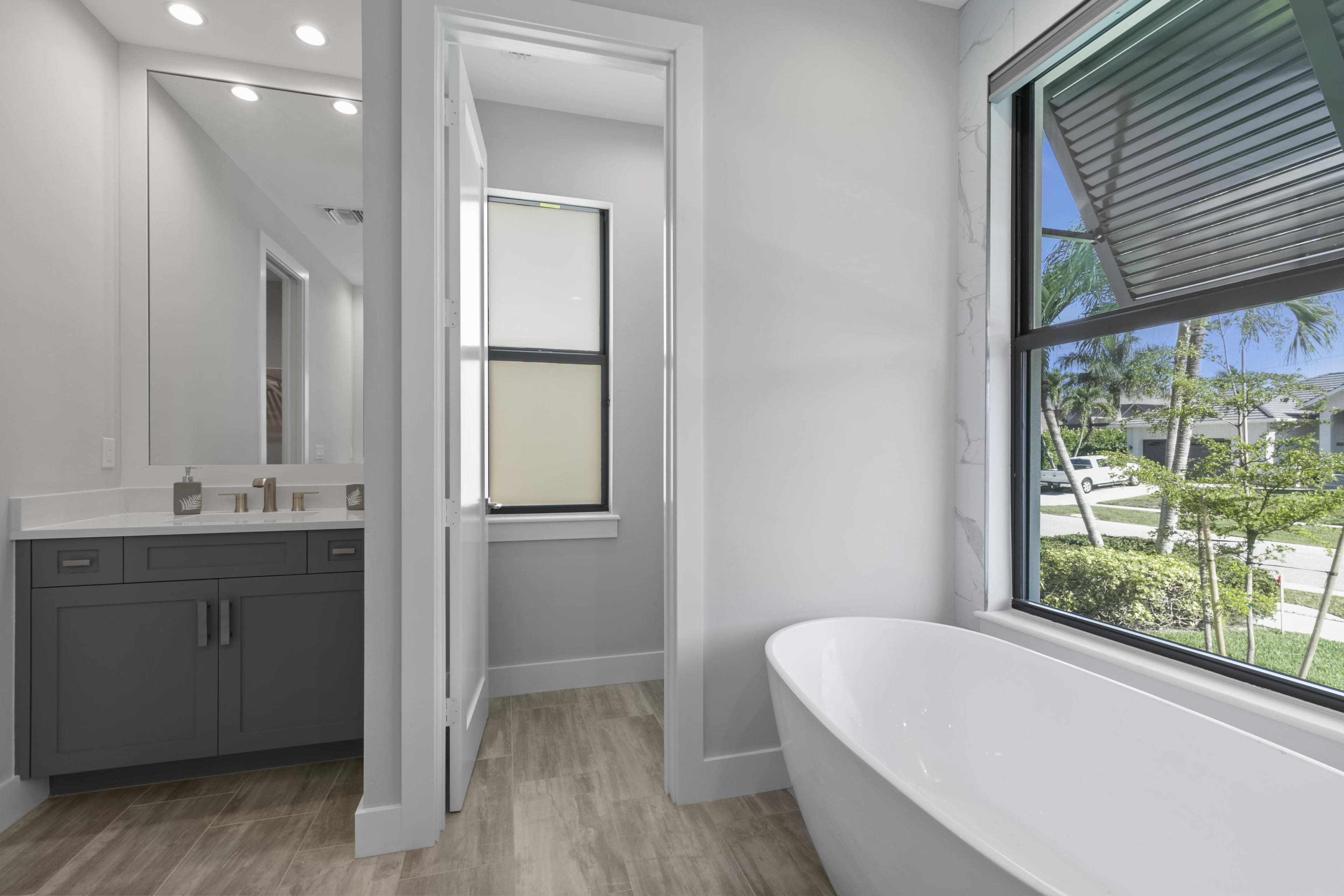 A modern bathroom features a freestanding tub, a sink vanity, and a view of greenery outside through a large window.