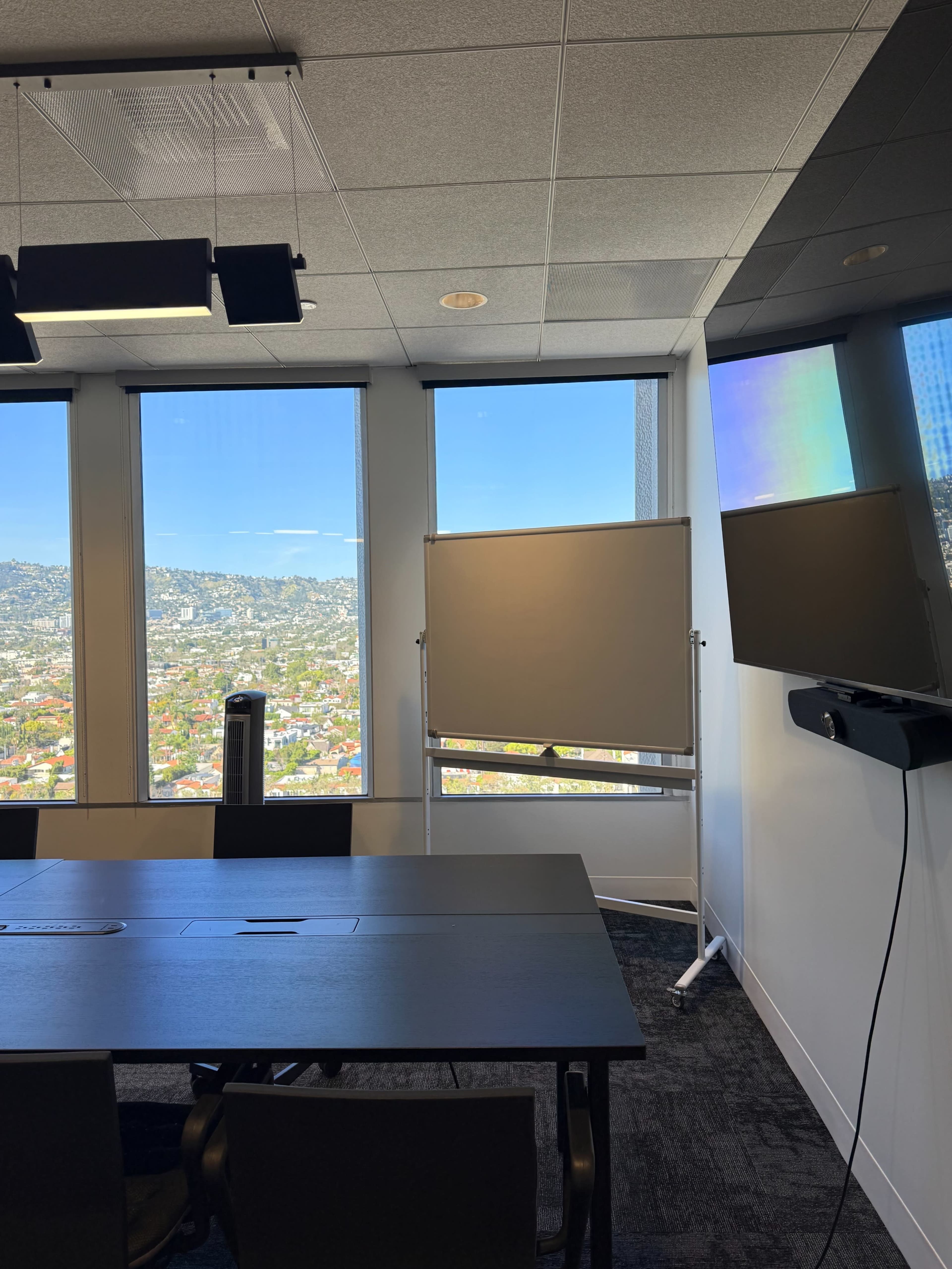 The image shows a modern conference room with a large table, a whiteboard, and two screens, overlooking a cityscape through large windows.