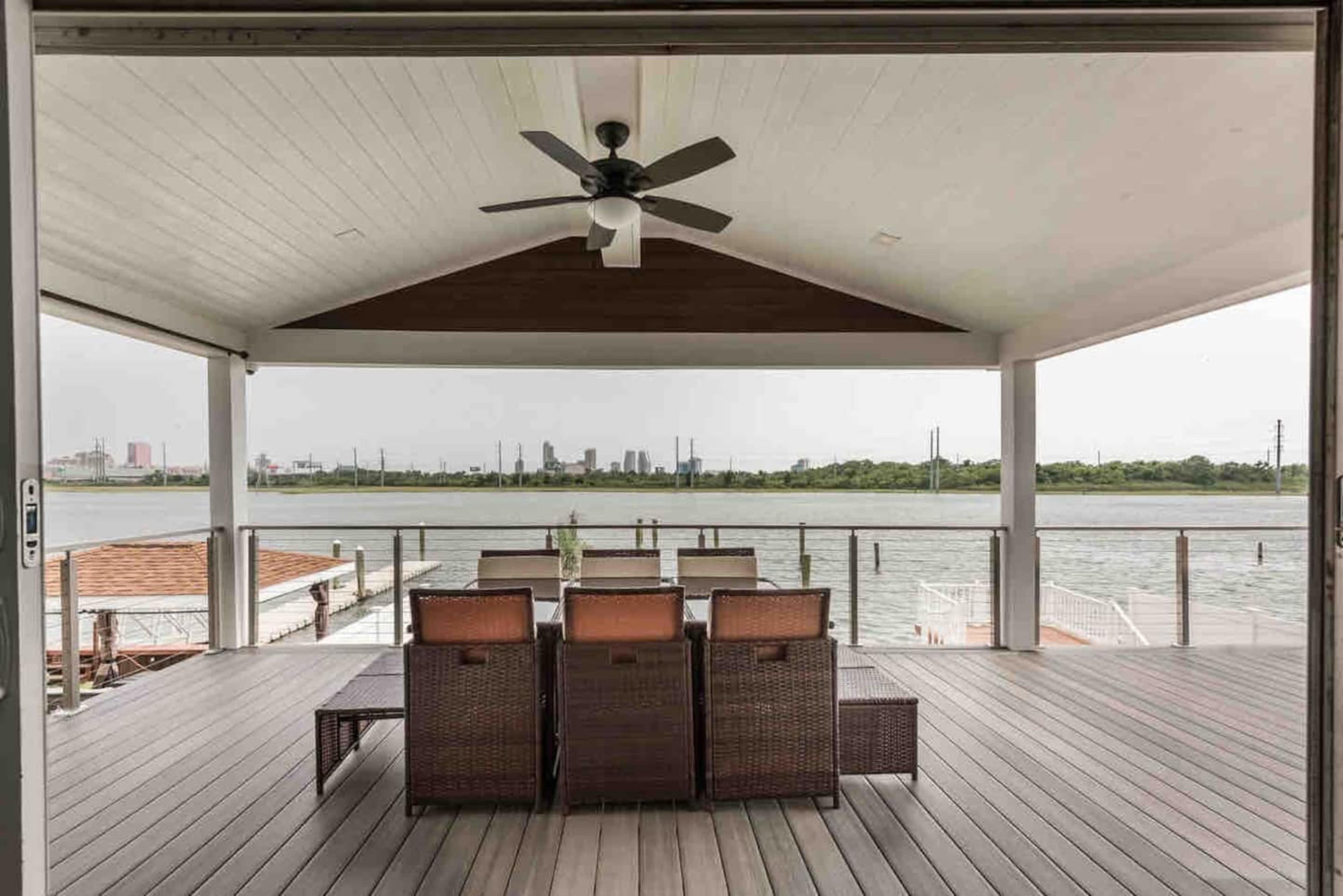 A covered porch features a dining table with chairs facing a body of water and a city skyline in the background.