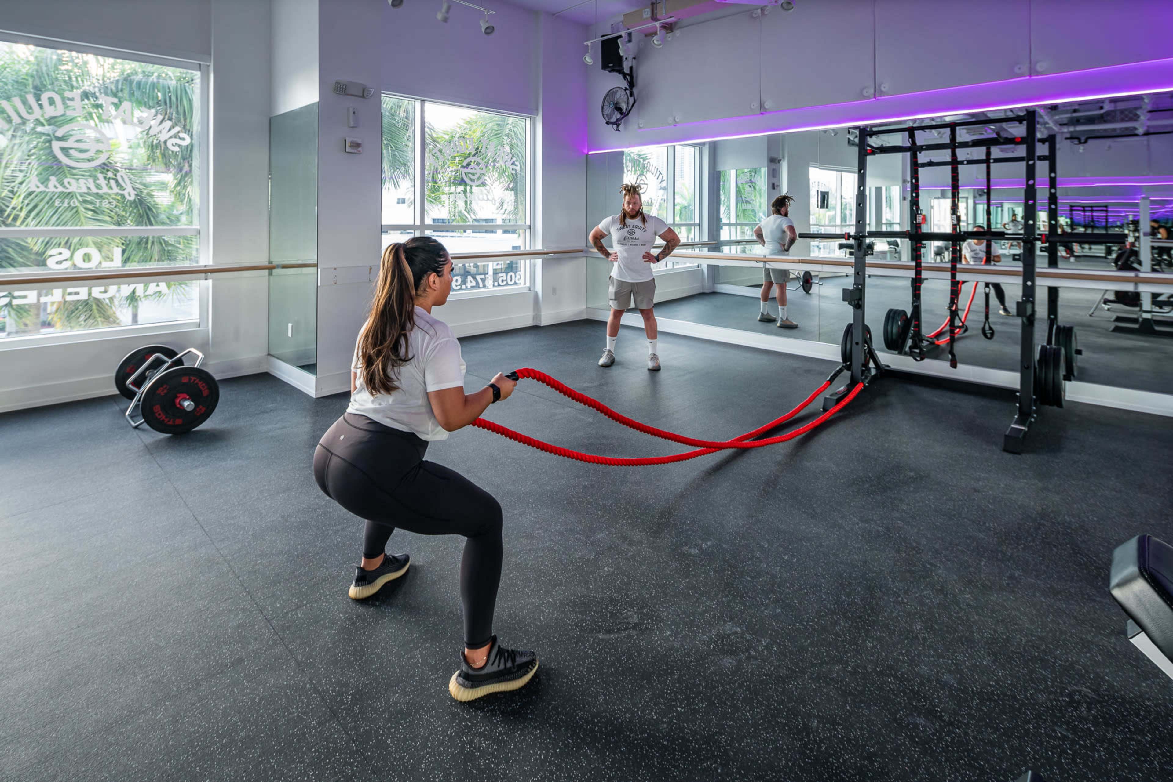 A woman performs a battle rope exercise while a trainer observes in a modern gym space.