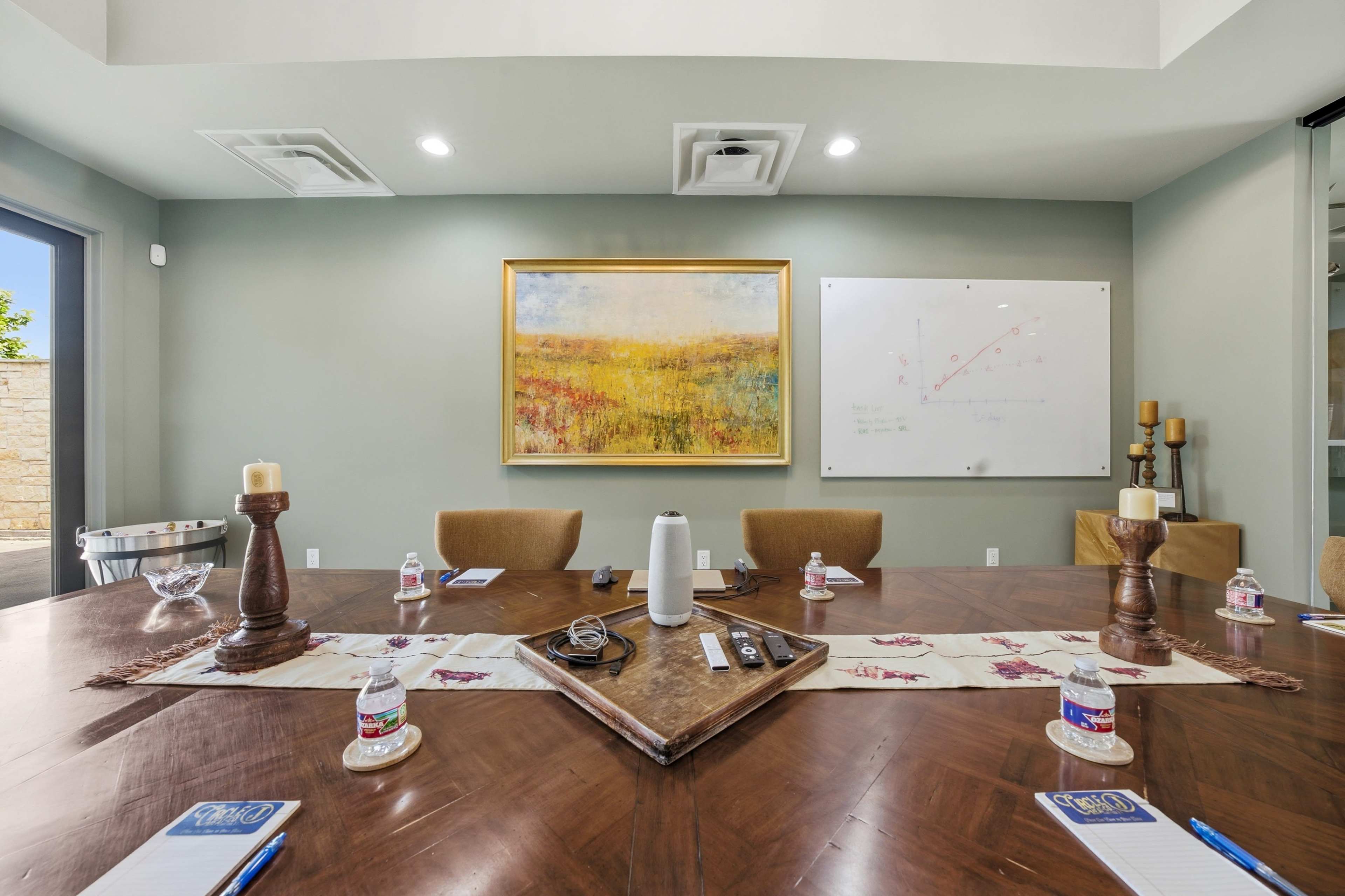 A modern conference room features a large wooden table set with notepads and water bottles, surrounded by chairs, with an abstract painting and presentation board on the walls.