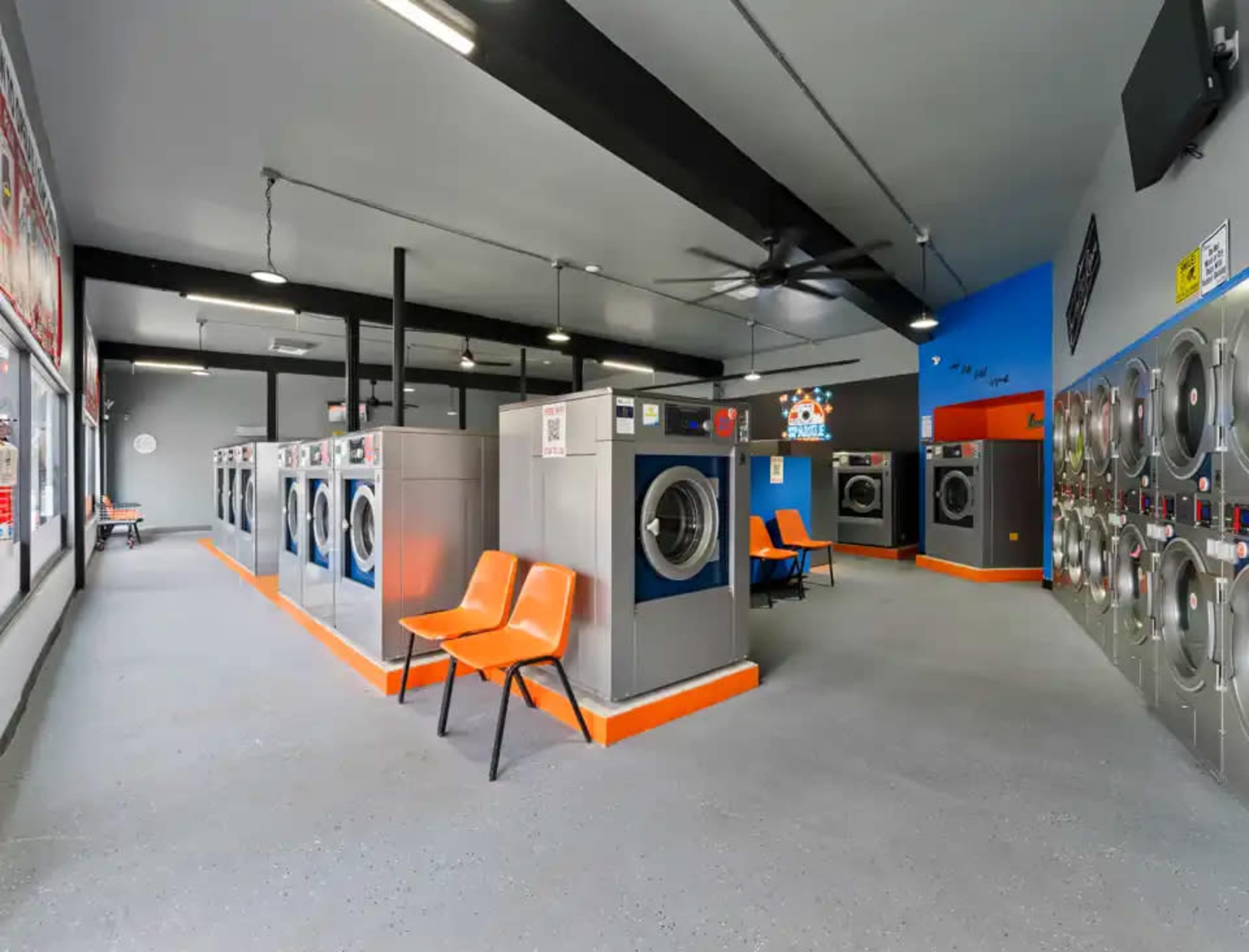 The image shows an empty laundromat featuring rows of washing machines and dryers, with orange chairs positioned nearby.