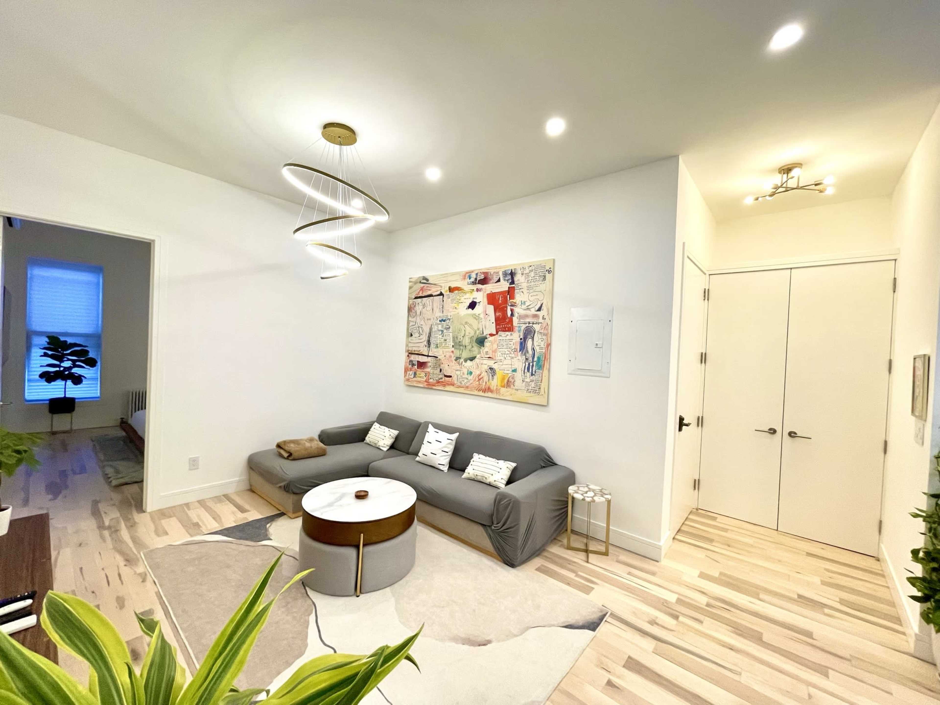 The image shows a modern living room featuring a gray sectional sofa, a round coffee table, and a large piece of abstract artwork on the wall, with a bright overhead light and plants in the foreground.