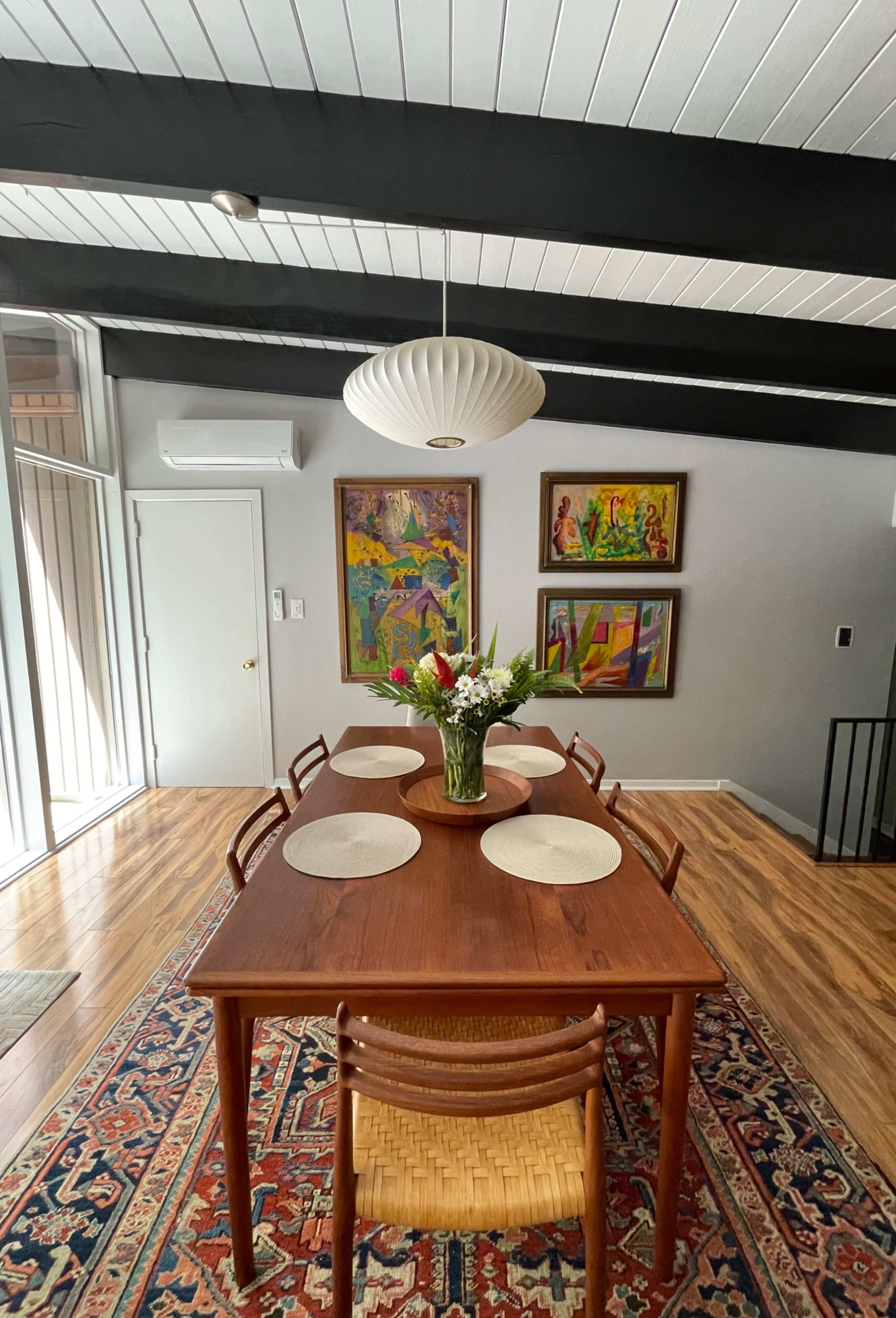 A wooden dining table with a flower arrangement and placemats is centrally located in a room featuring two colorful paintings and exposed beam ceilings.