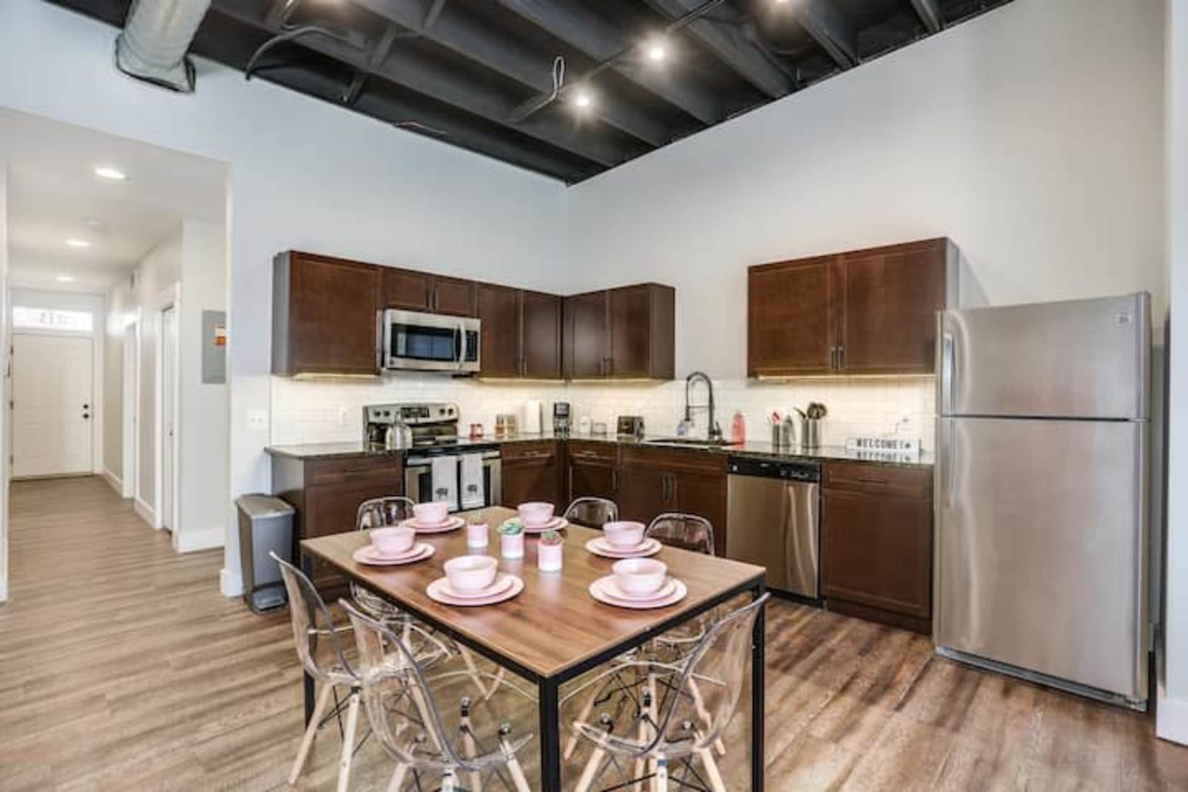 A modern kitchen with dark wooden cabinets, stainless steel appliances, and a dining table set with pink plates and cups.