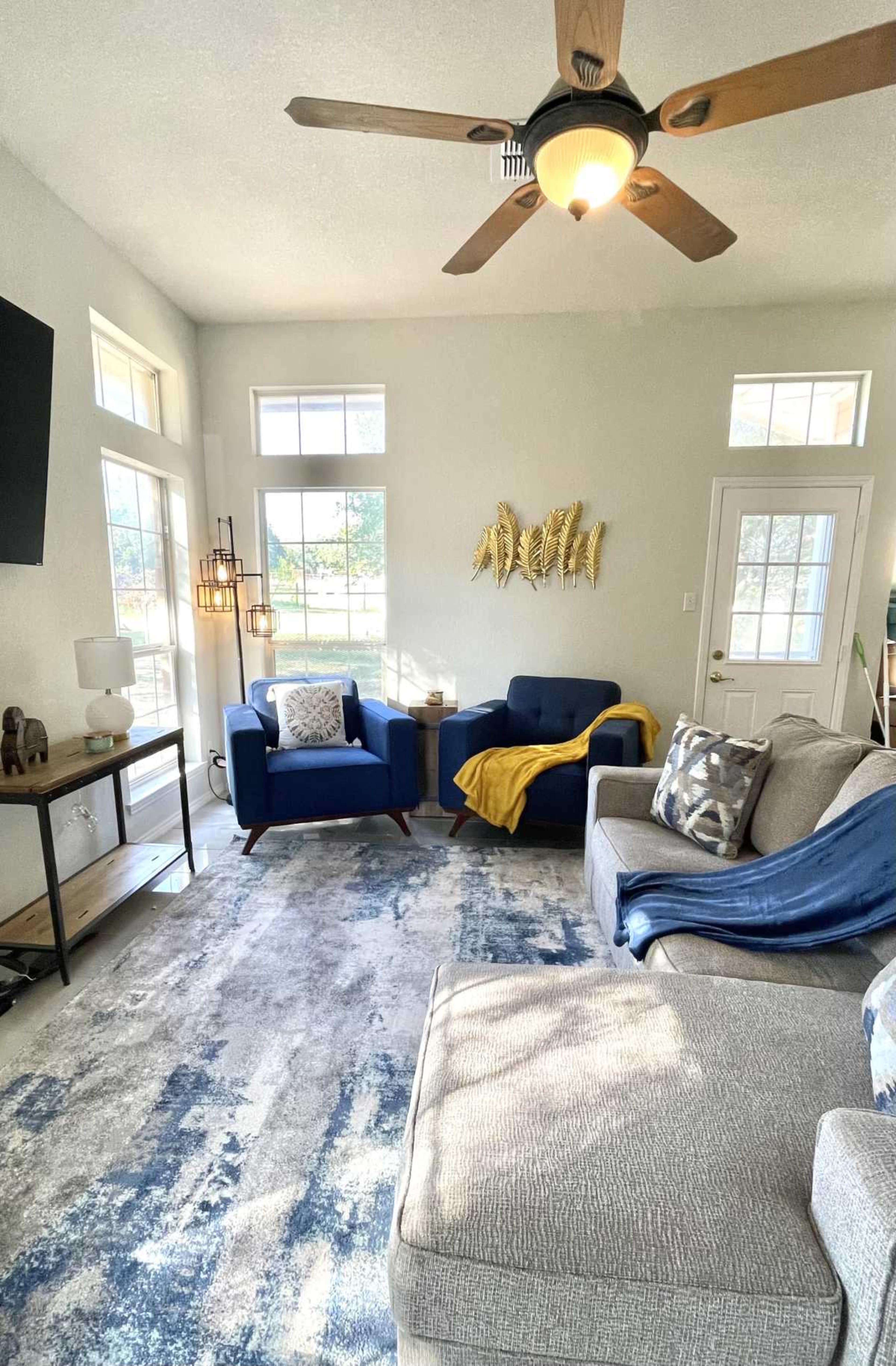 The living room features two blue chairs, a gray couch, a television, and a decorative wall installation, with large windows allowing natural light.