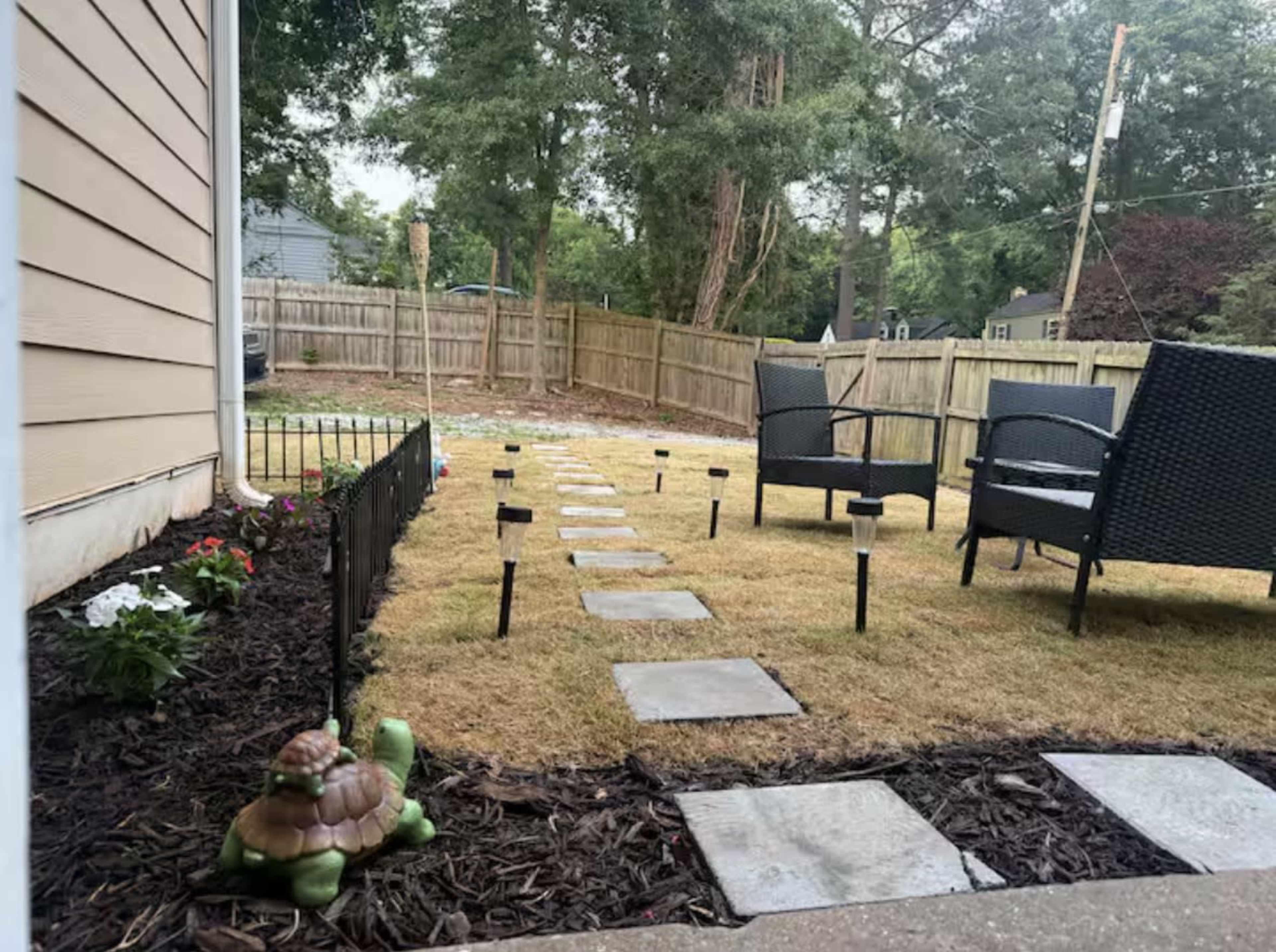 A pathway of stone slabs leads through a grassy area bordered by flower beds and solar lights, with two chairs sitting nearby.