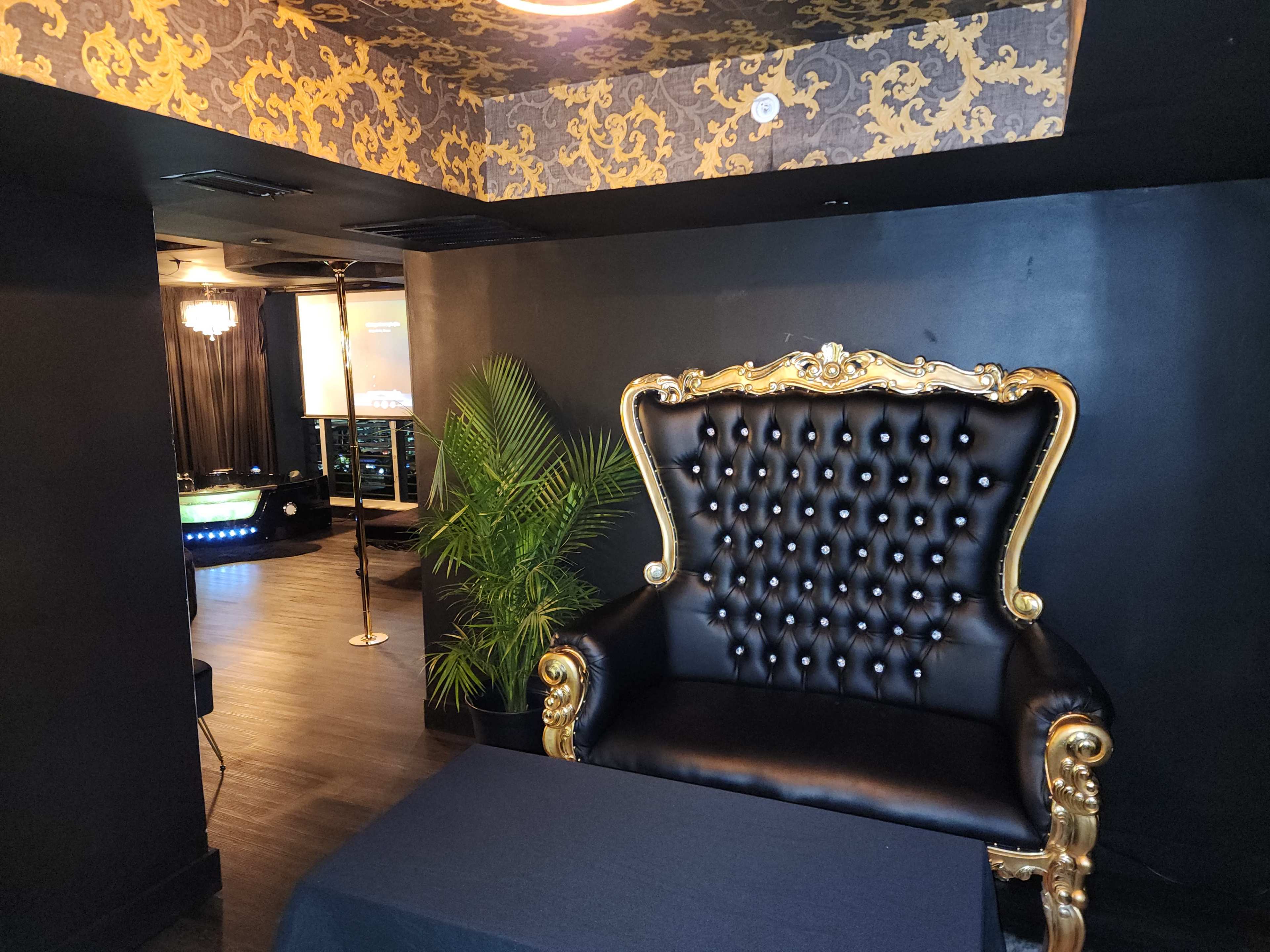 The image shows a lavish black and gold decorated room featuring an ornate golden throne chair in the foreground, with a table and a green plant beside it, and a projector screen visible in the background.