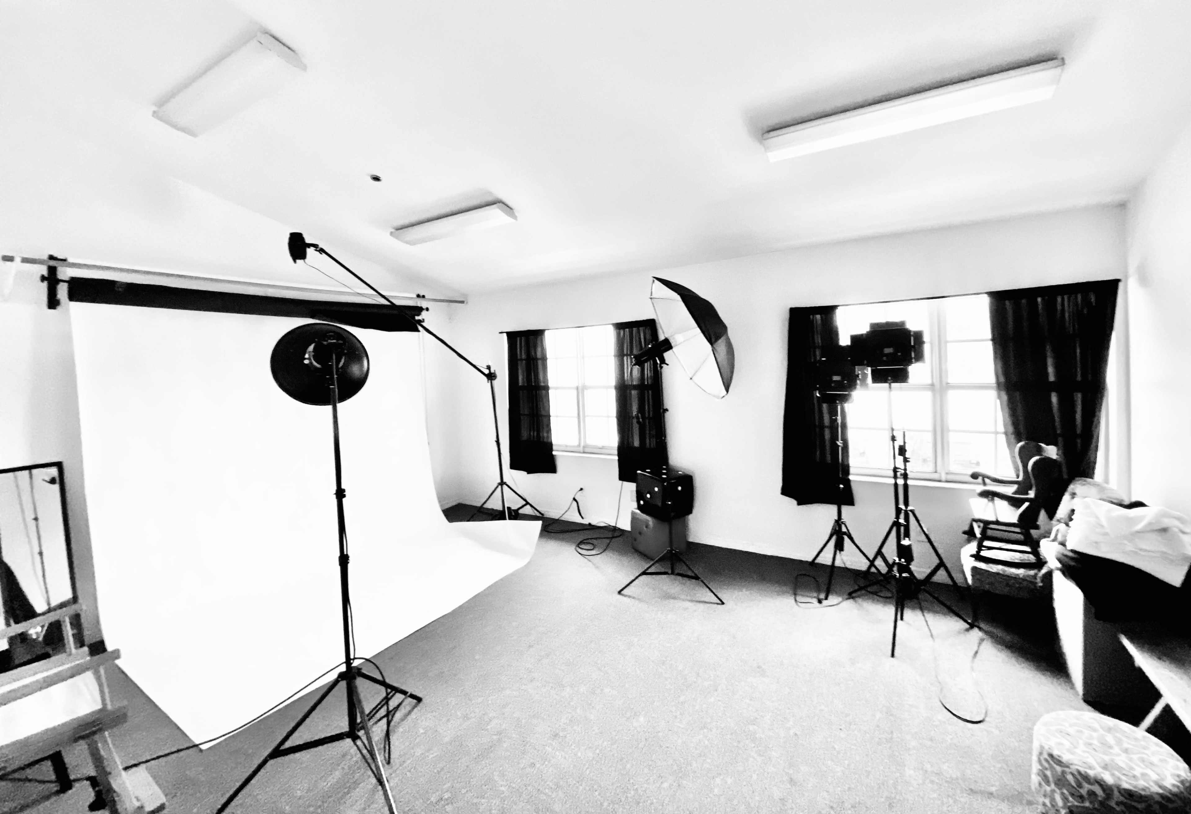 The image shows a photography studio with a white backdrop, lighting equipment, and a chair positioned in front of the window.