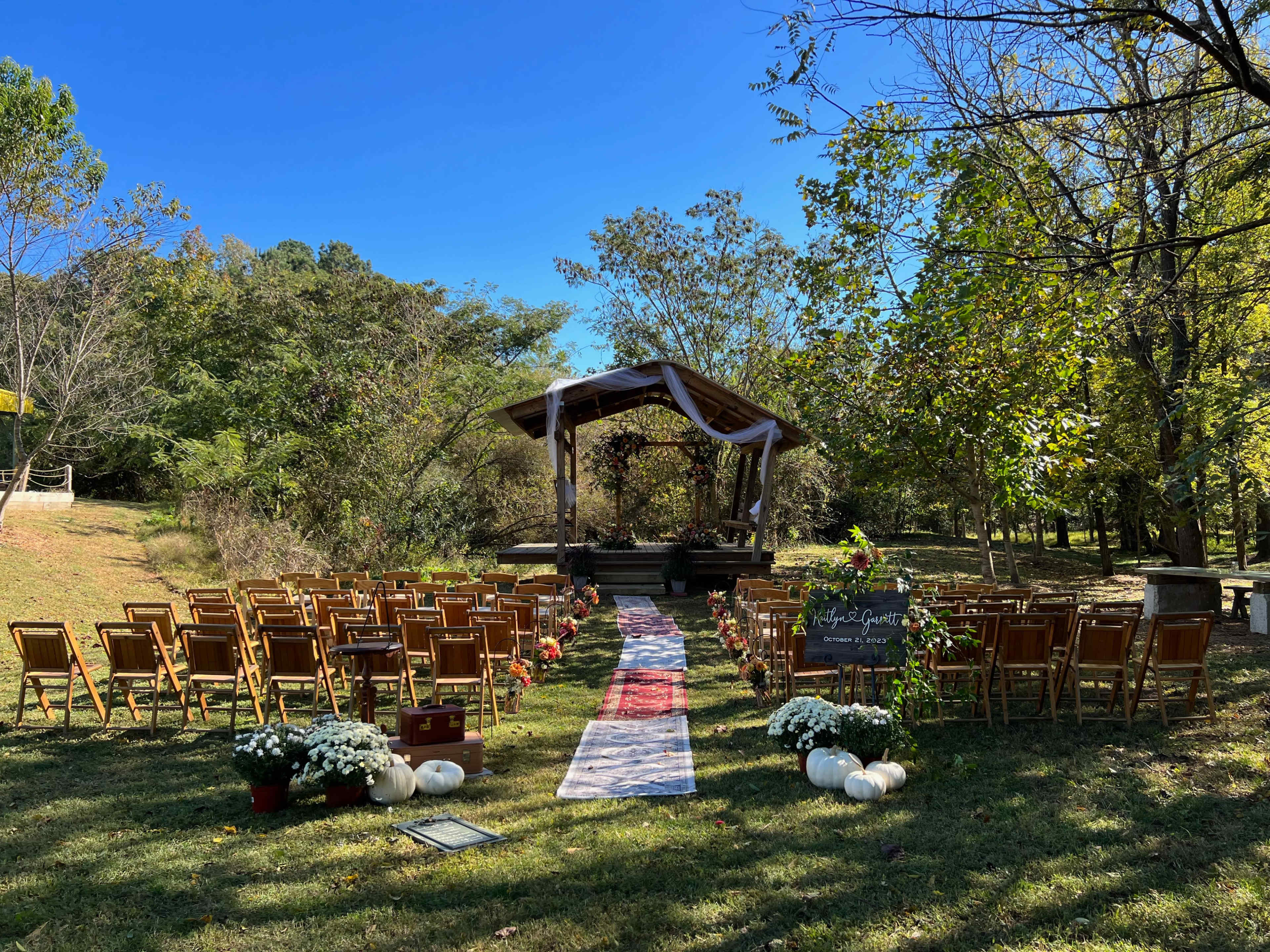 A wedding ceremony setup features rows of wooden chairs arranged on a grassy area leading to a decorated altar under a gazebo, surrounded by trees.