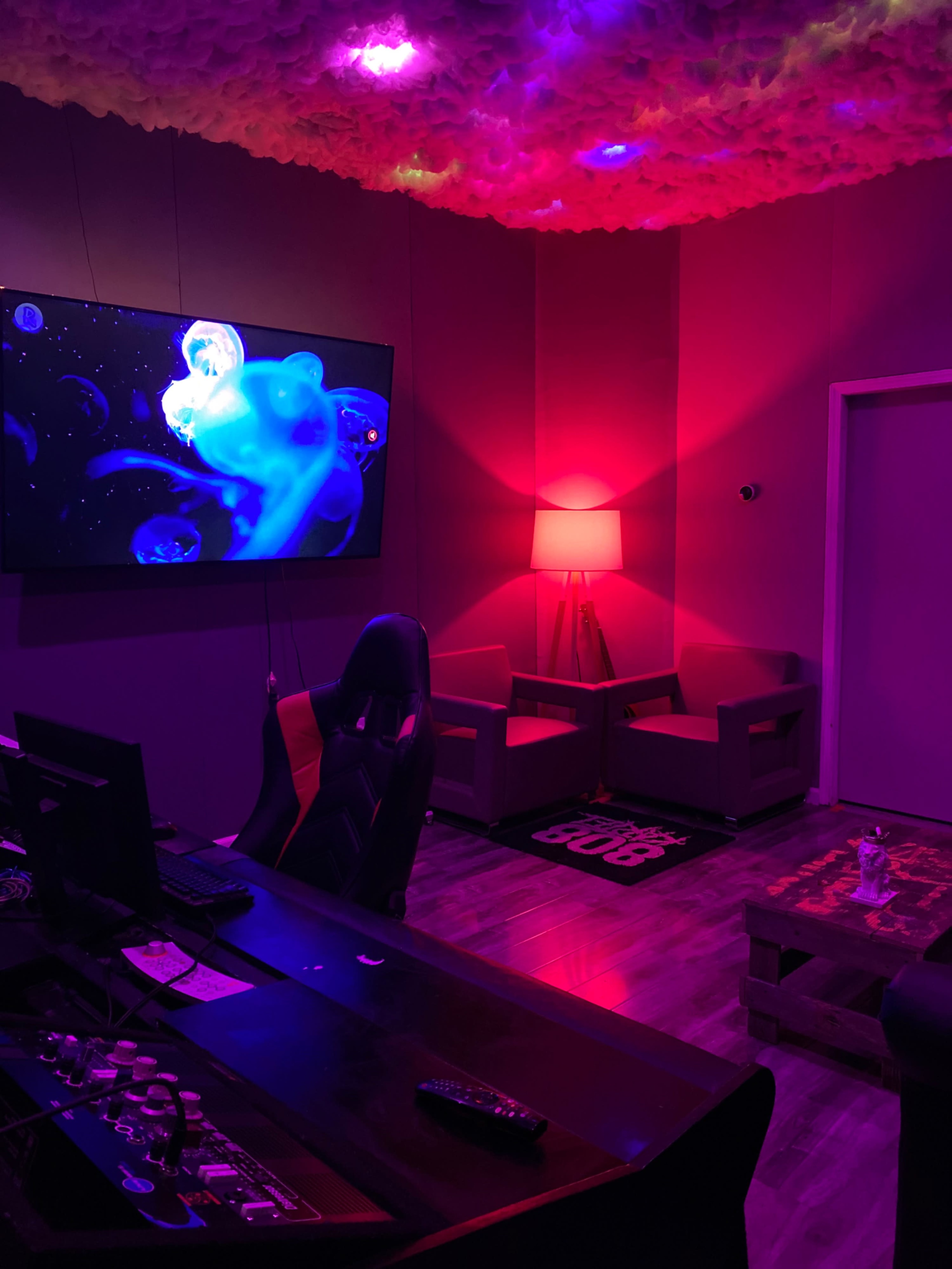A dimly lit room with a large wall-mounted screen displaying colorful visuals, a desk with a computer, and two chairs beside a lamp emitting pink light.