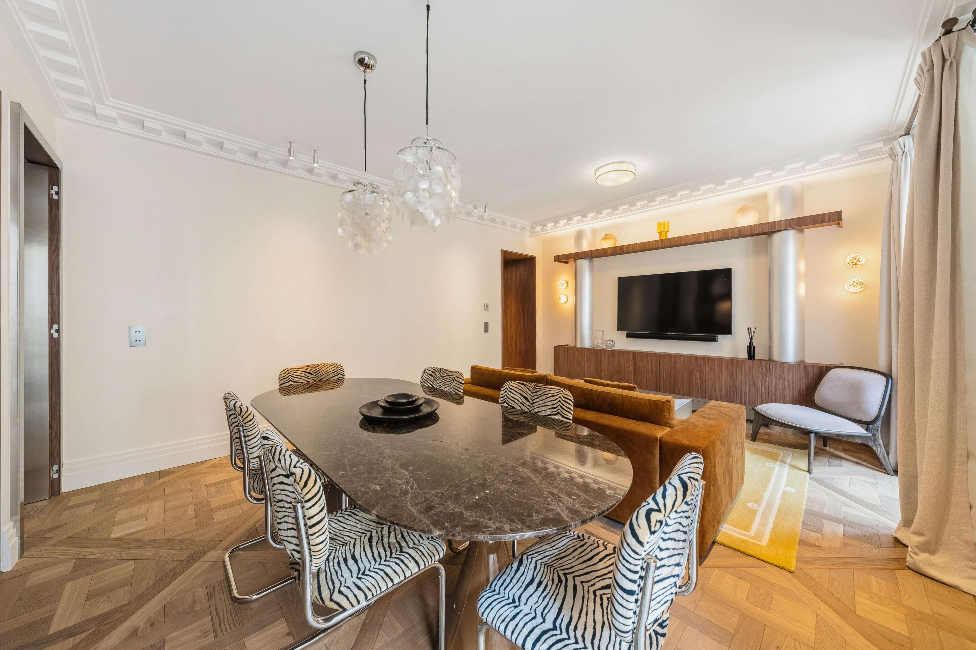 The image depicts a modern dining area featuring a dark marble table with striped chairs, a cozy living space with a brown sofa, and a wall-mounted television.