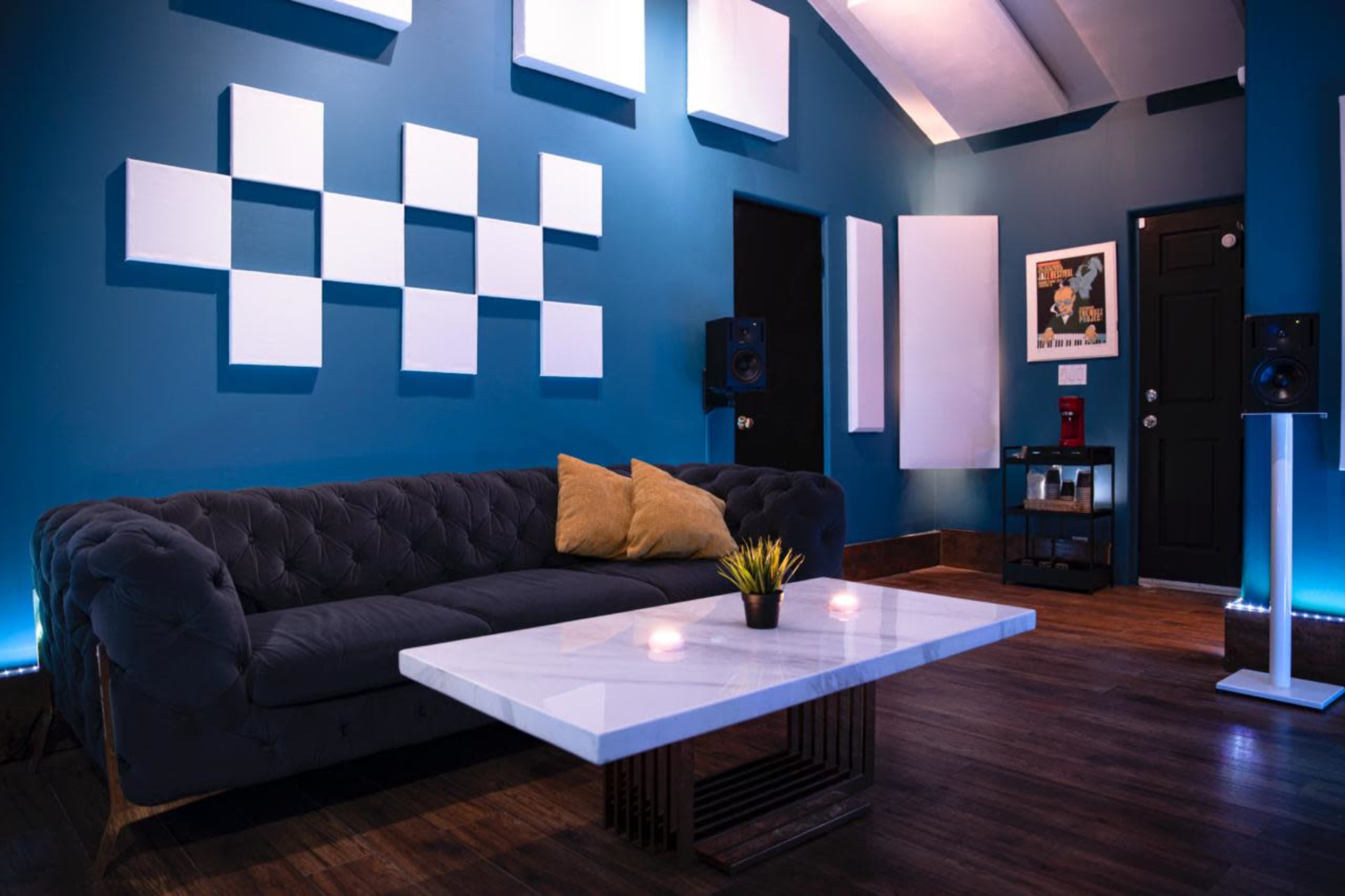 A modern lounge area features a dark couch with two decorative pillows, a marble coffee table, and acoustic panels on the blue walls, illuminated by ambient lighting.