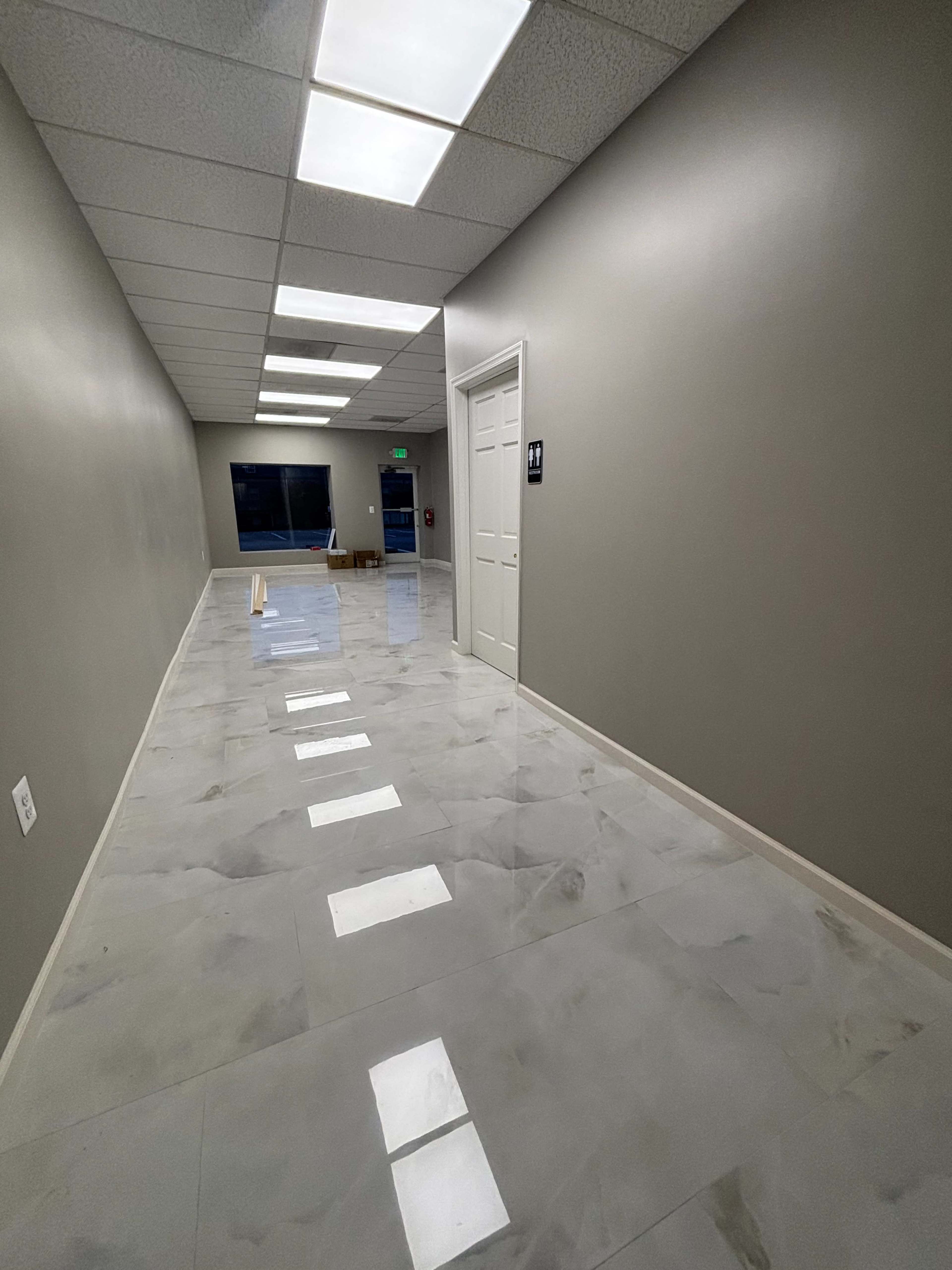 The image shows a long, well-lit corridor with grey walls and shiny tiled flooring, leading to a door on the right and windows at the end.