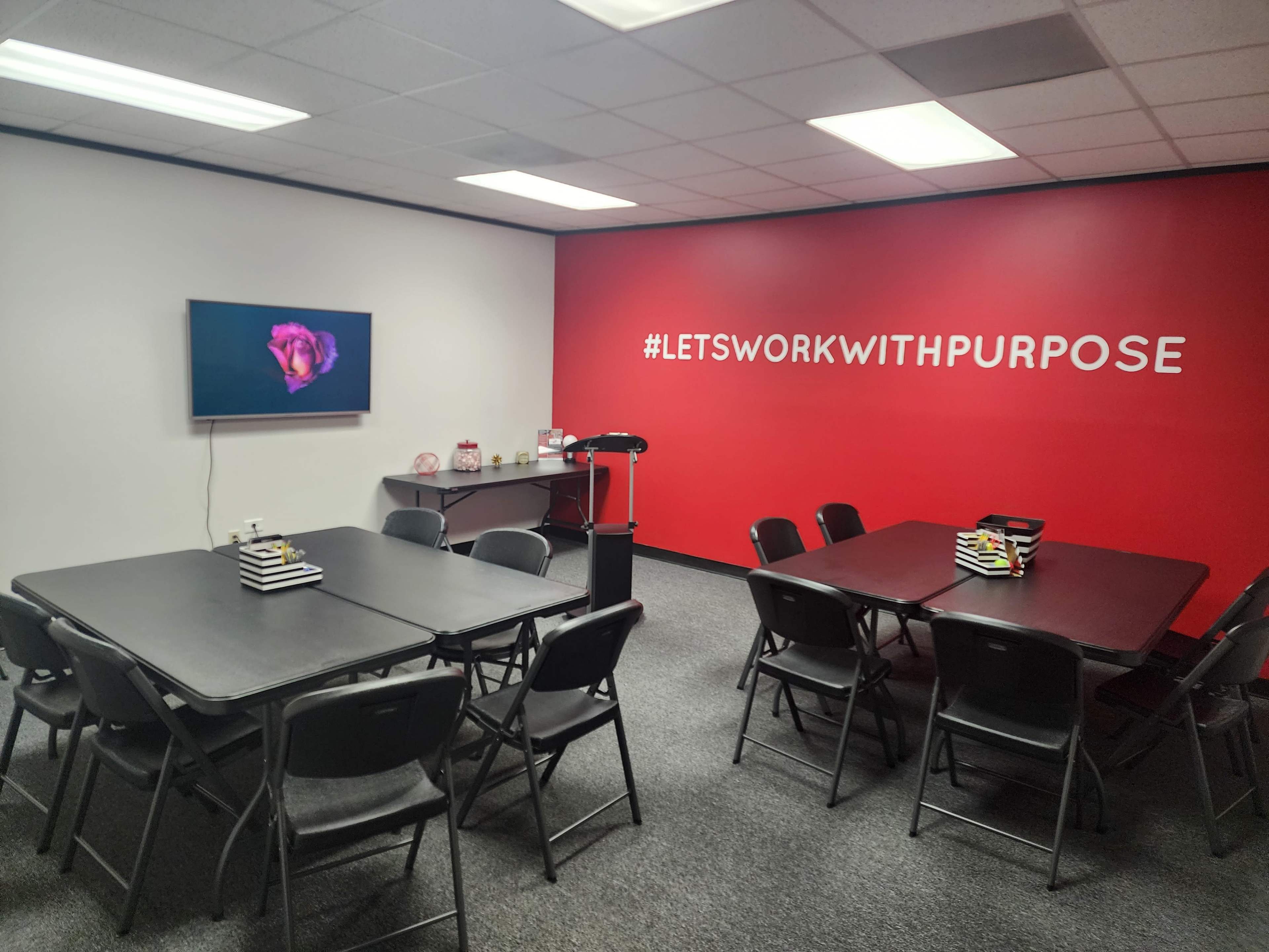 The image shows a meeting room with several tables and chairs arranged, a television displaying a floral image, and a wall featuring the phrase "#LET'SWORKWITHPURPOSE" in large letters.