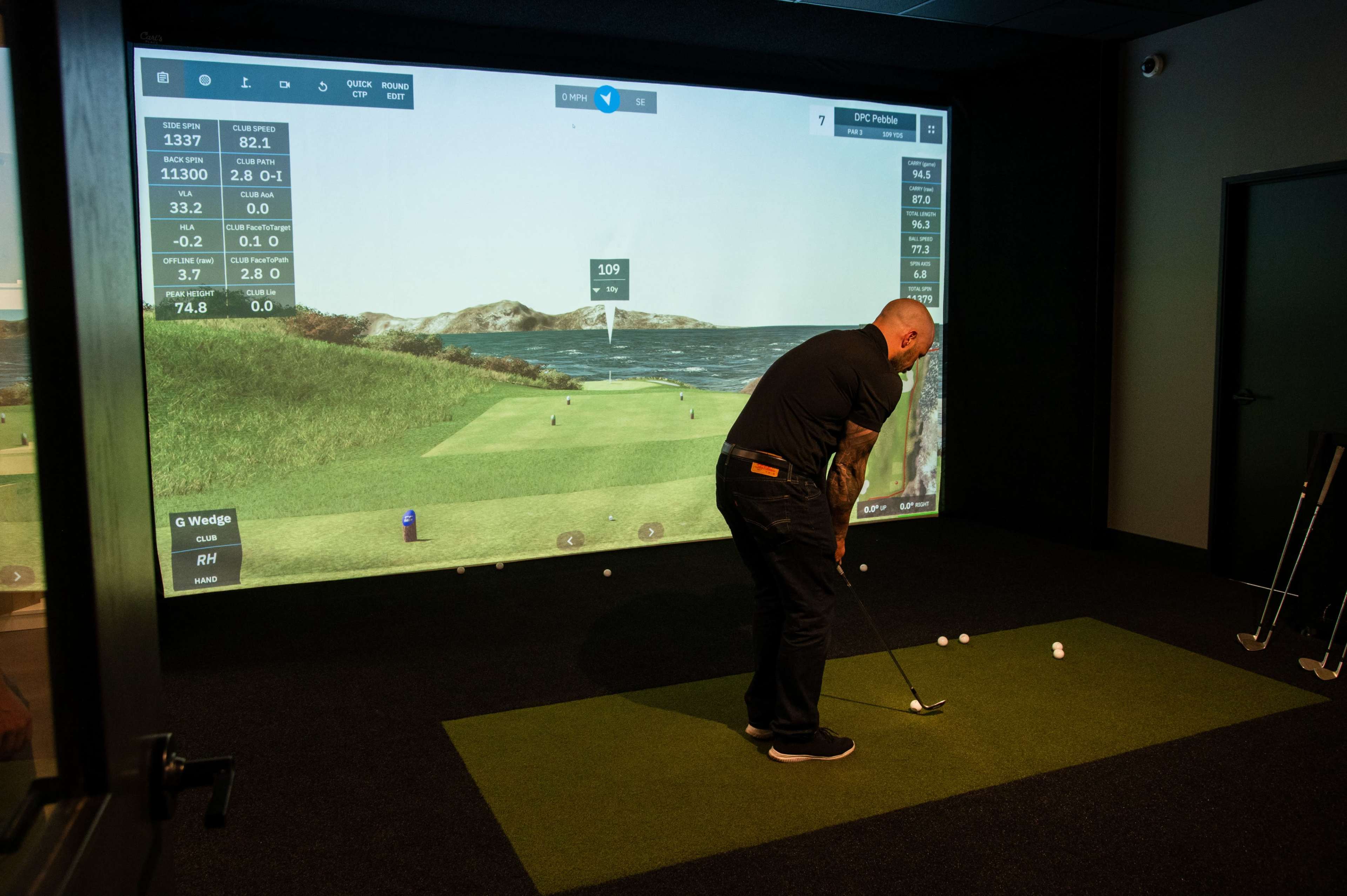 A person is taking a golf shot in an indoor simulator that displays a virtual golf course on a large screen.