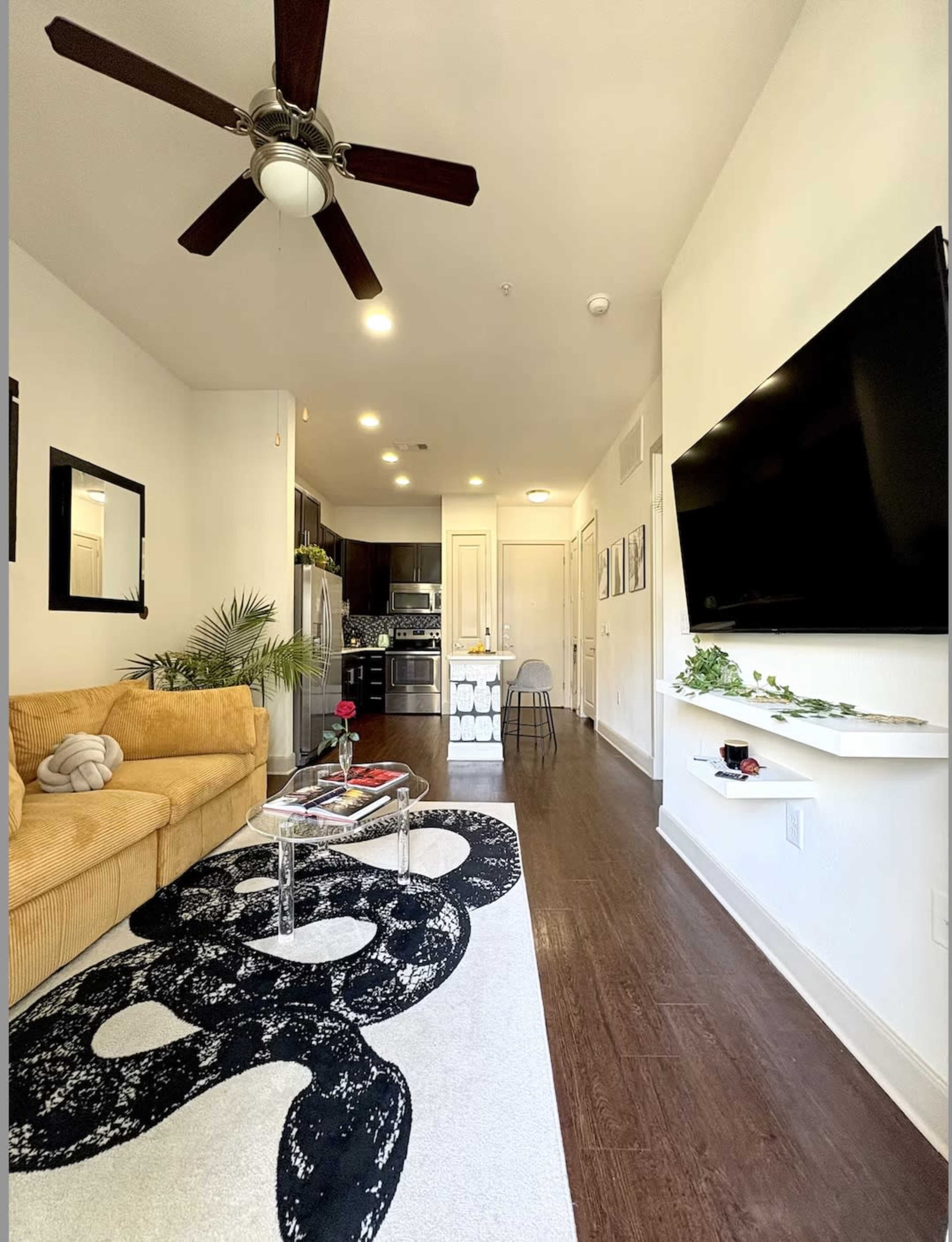 A modern living area features a yellow sofa, a black and white patterned area rug, and a wall-mounted television, with a kitchen visible in the background.