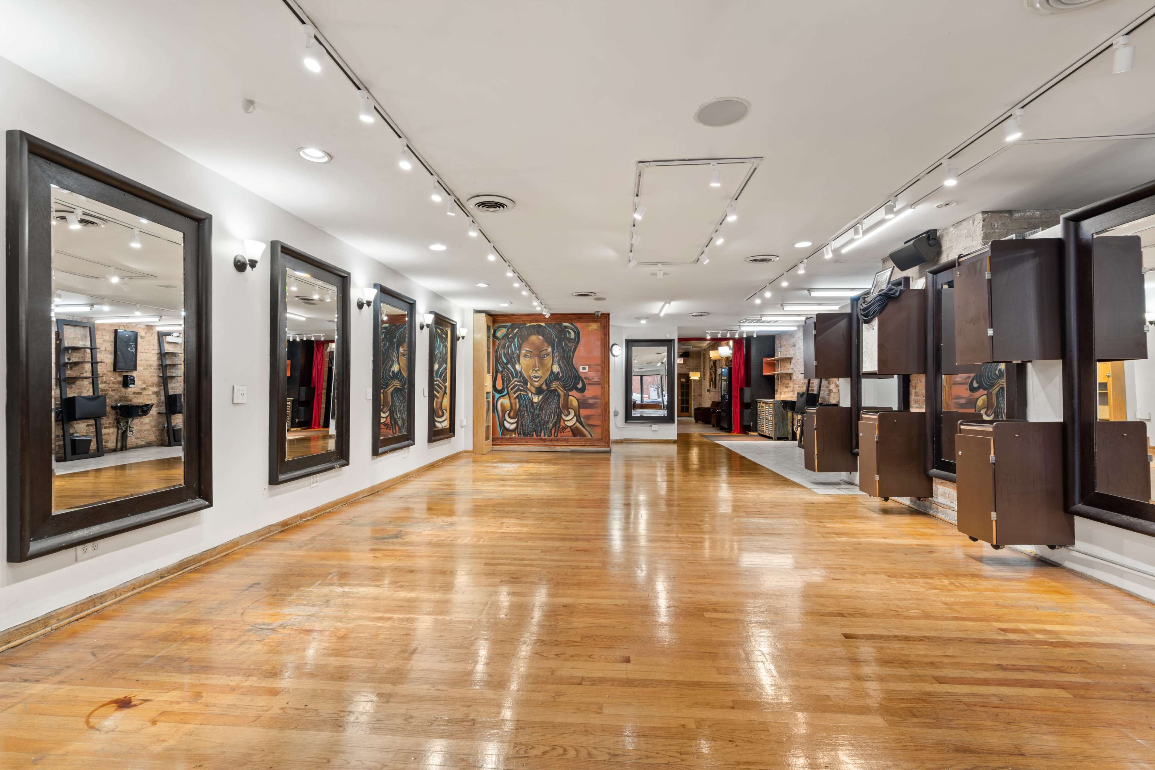 The image shows a spacious interior with polished wooden floors, featuring large mirrors on the walls and an art mural at one end.
