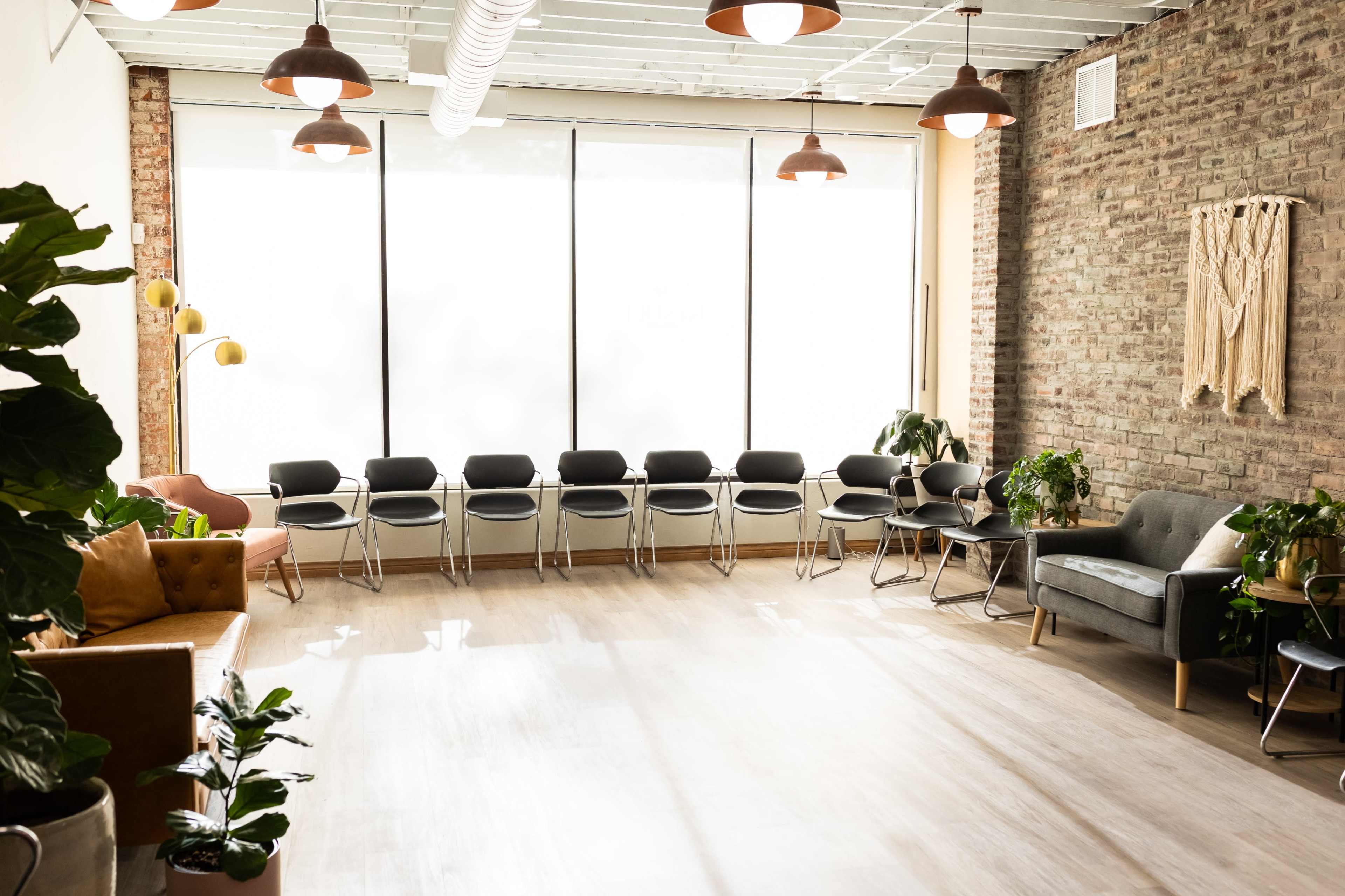 A spacious room features a row of black chairs along a large window, with plants and varied seating options arranged throughout.