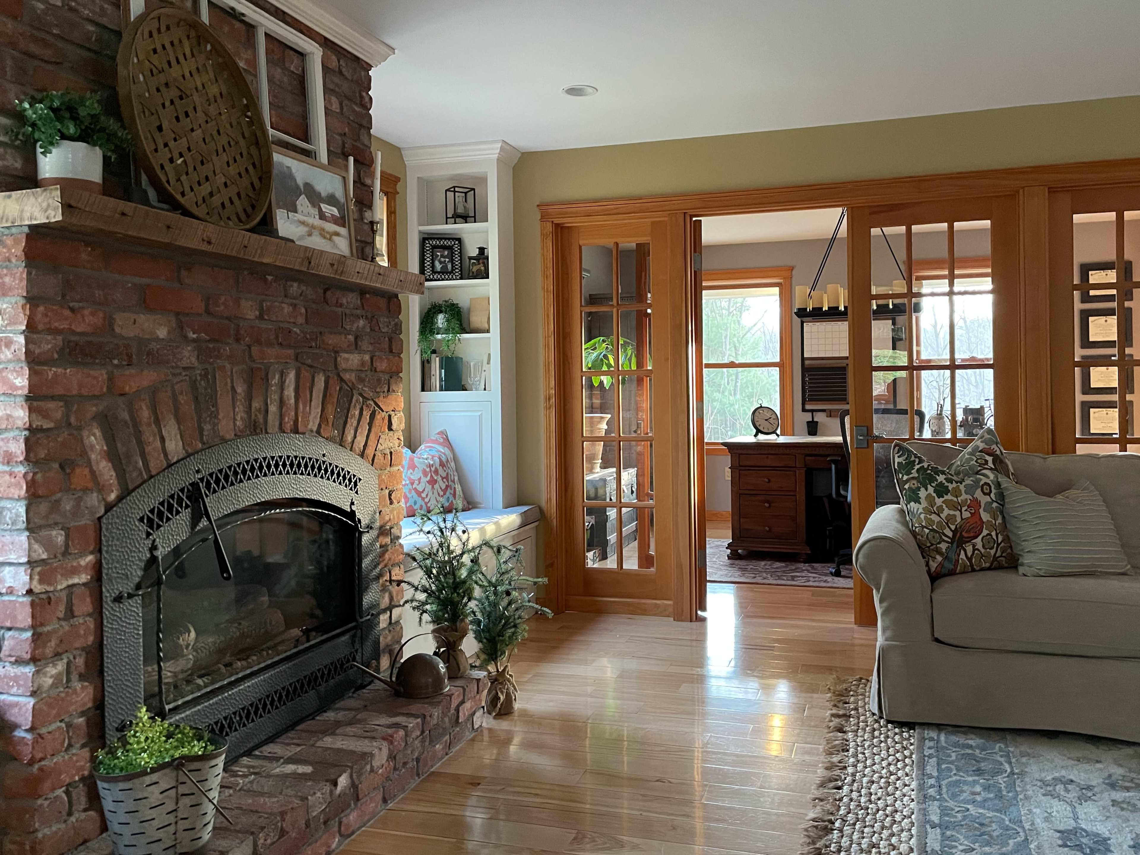 A cozy living room featuring a brick fireplace, a comfortable sofa, and an adjoining room with a desk and large windows.