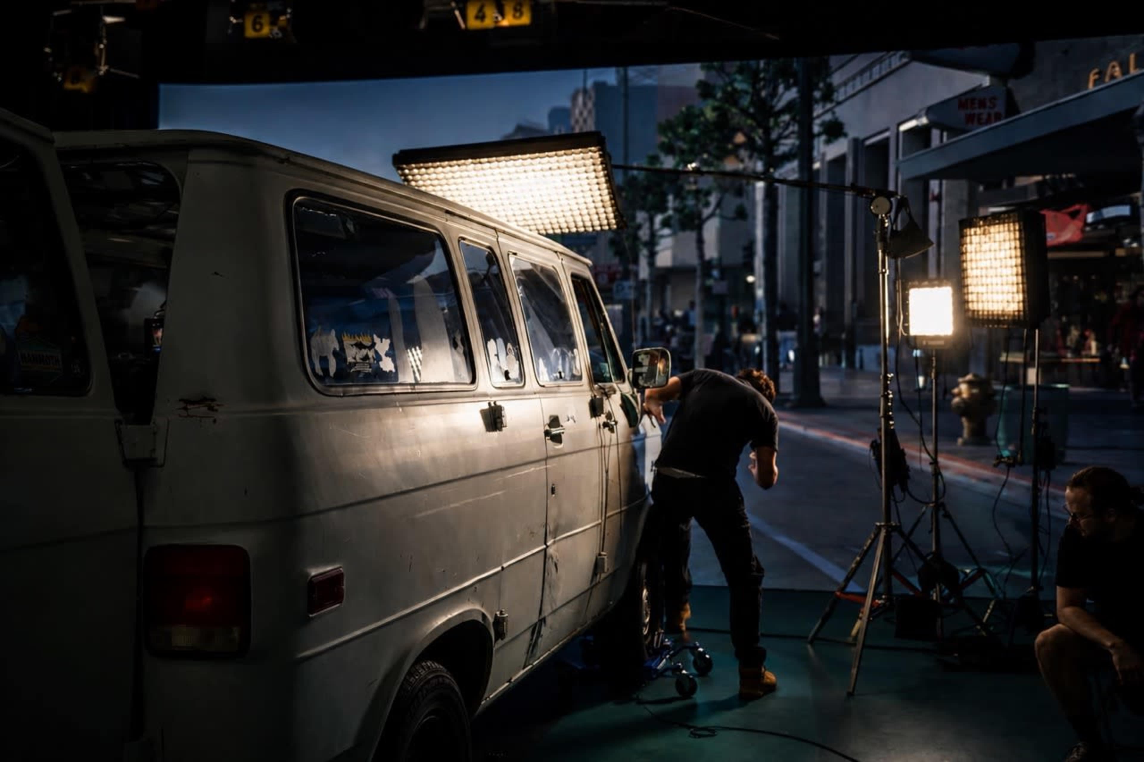 A crew member prepares equipment beside an old van under bright studio lights in an urban setting.