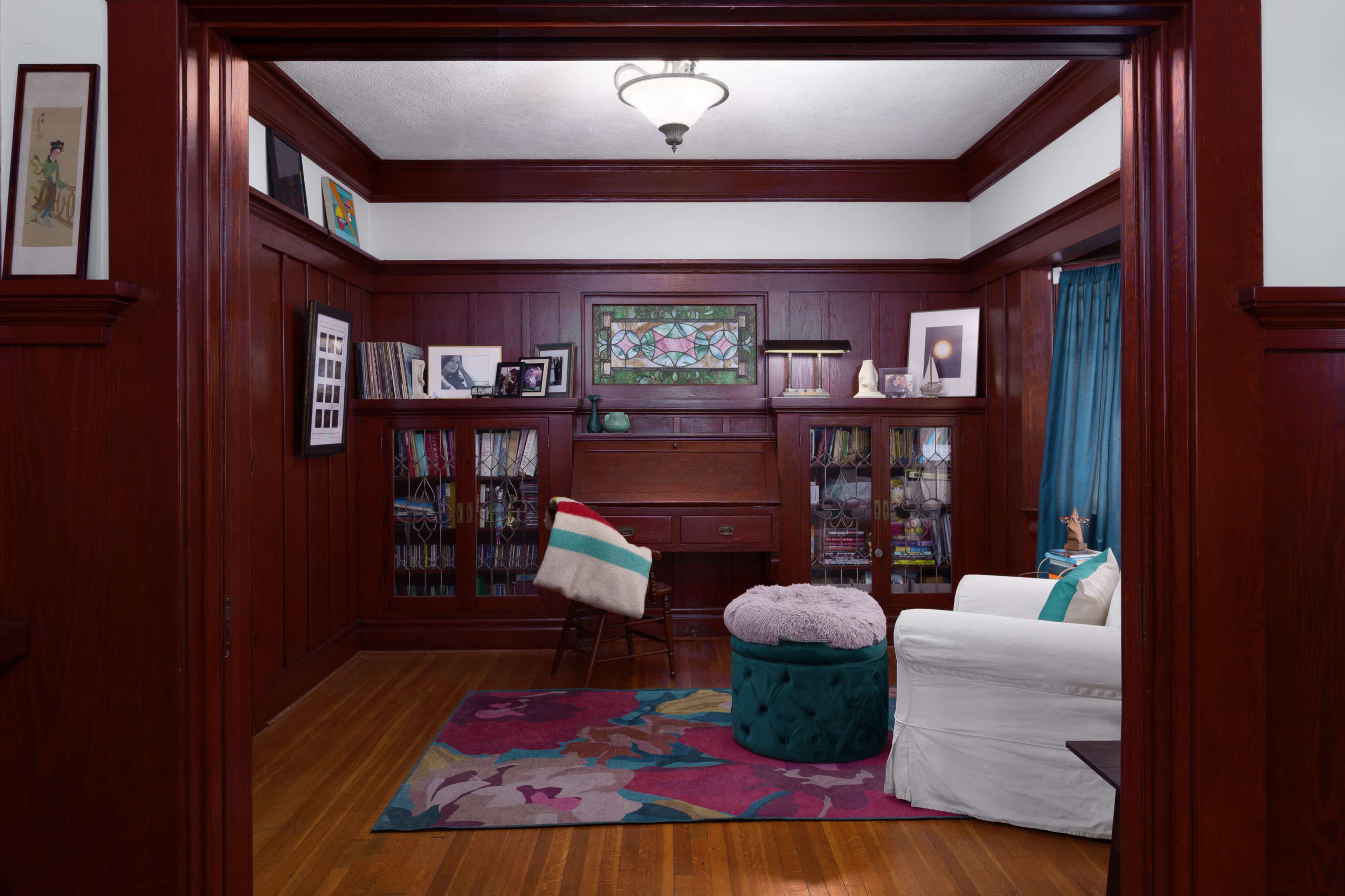 The image shows a cozy room with wooden walls, a colorful area rug, a white sofa, a green tufted ottoman, and shelves filled with books and decorative items.