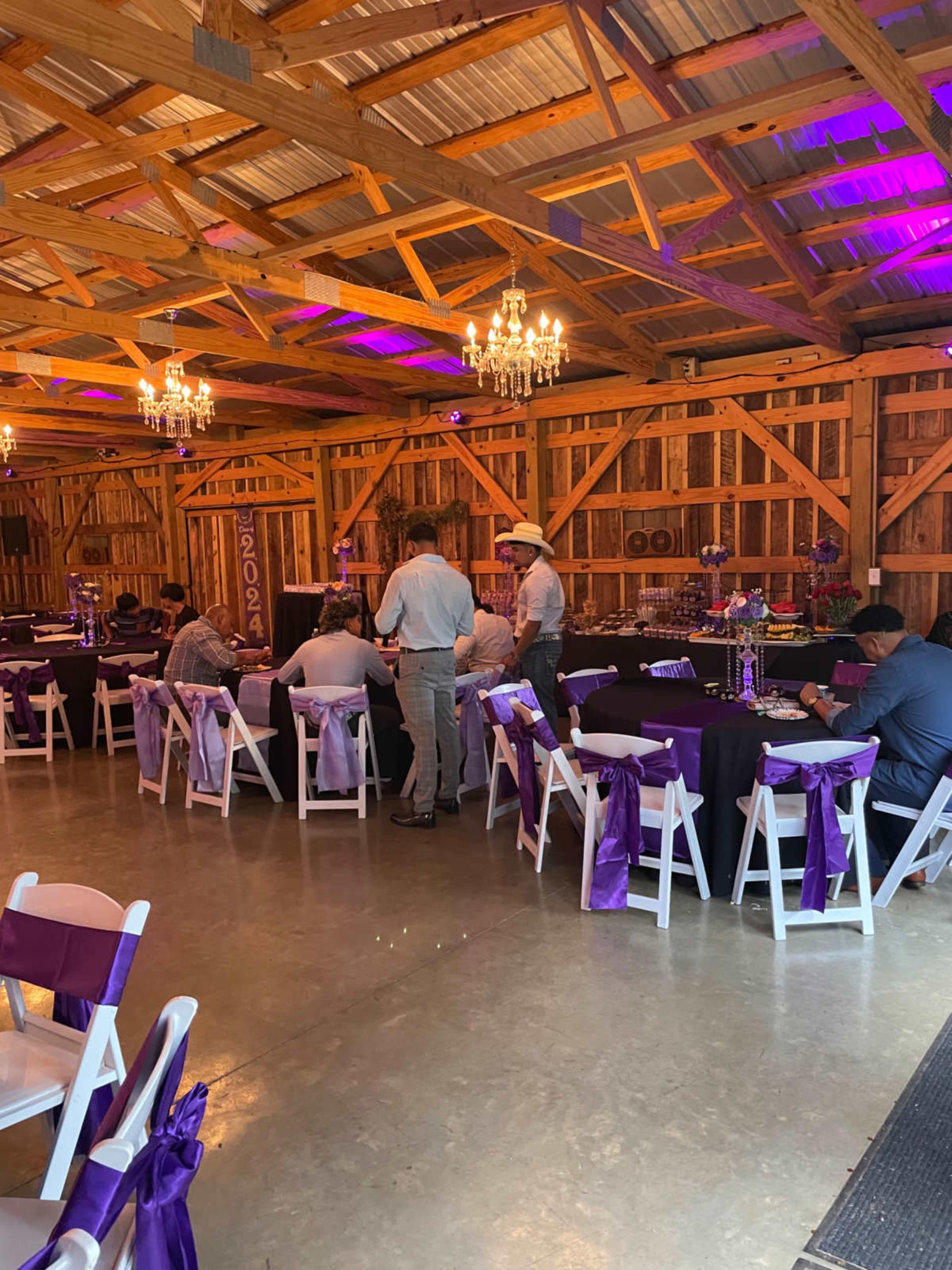 A decorated barn interior features rows of tables with purple tablecloths and chairs, illuminated by chandeliers and colorful lights.
