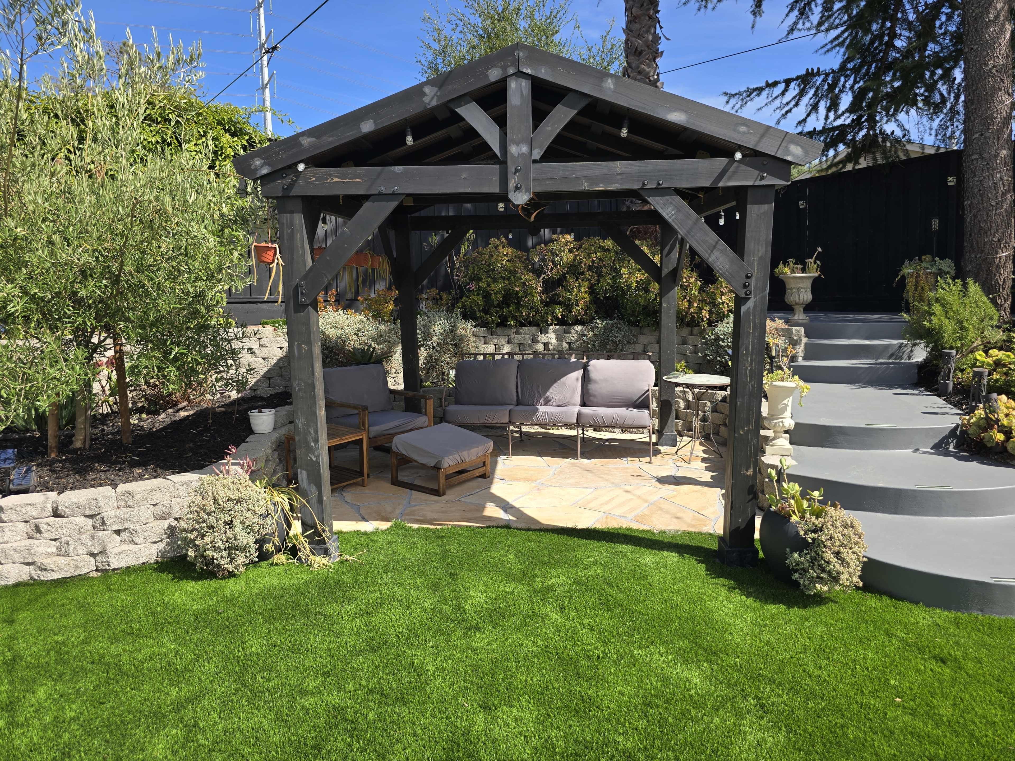 The image shows a shaded outdoor seating area with a wooden gazebo, surrounded by lush greenery and stone landscaping.