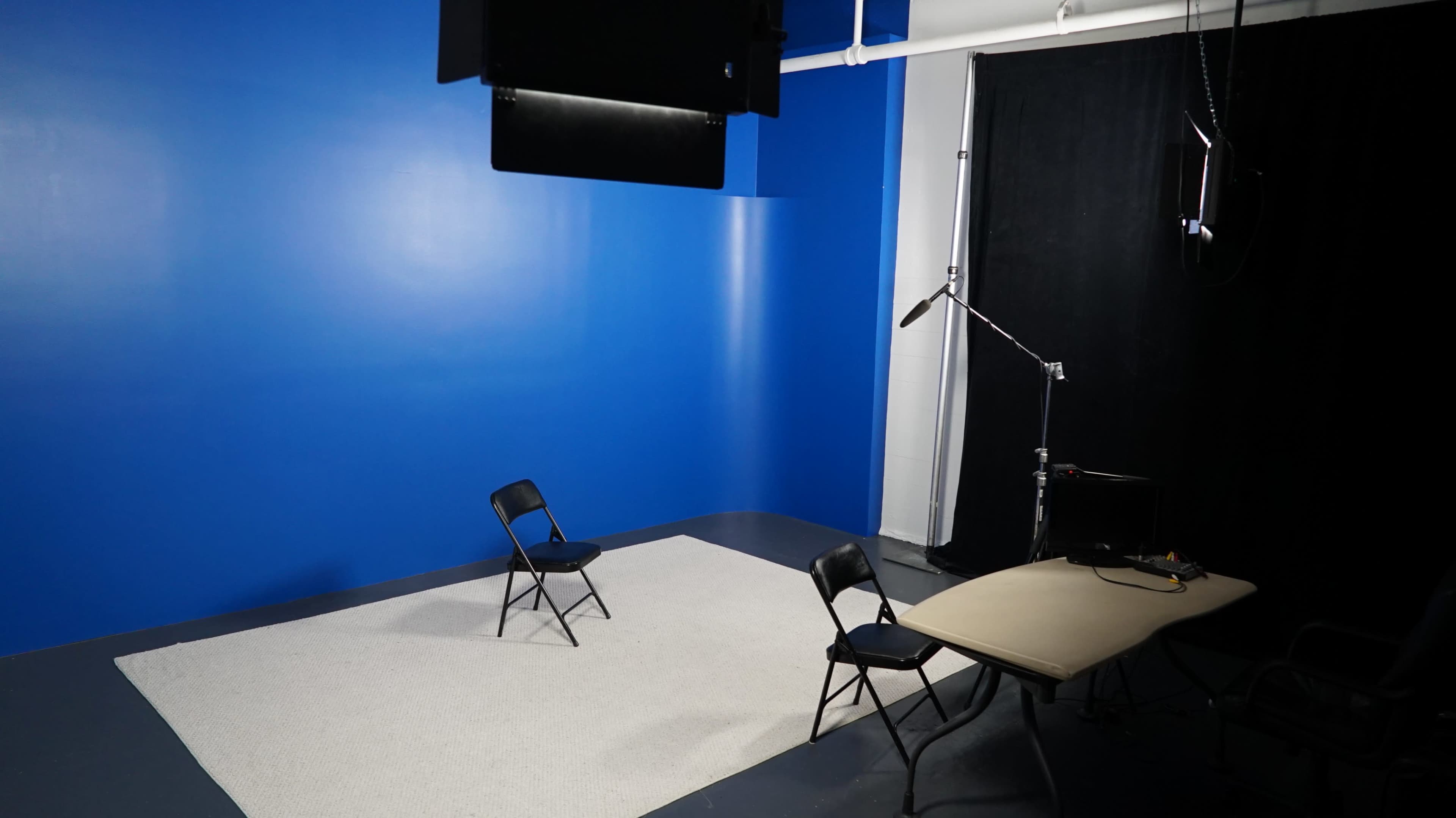 The image shows an empty studio space with a blue wall, two black chairs, and a table under soft lighting.