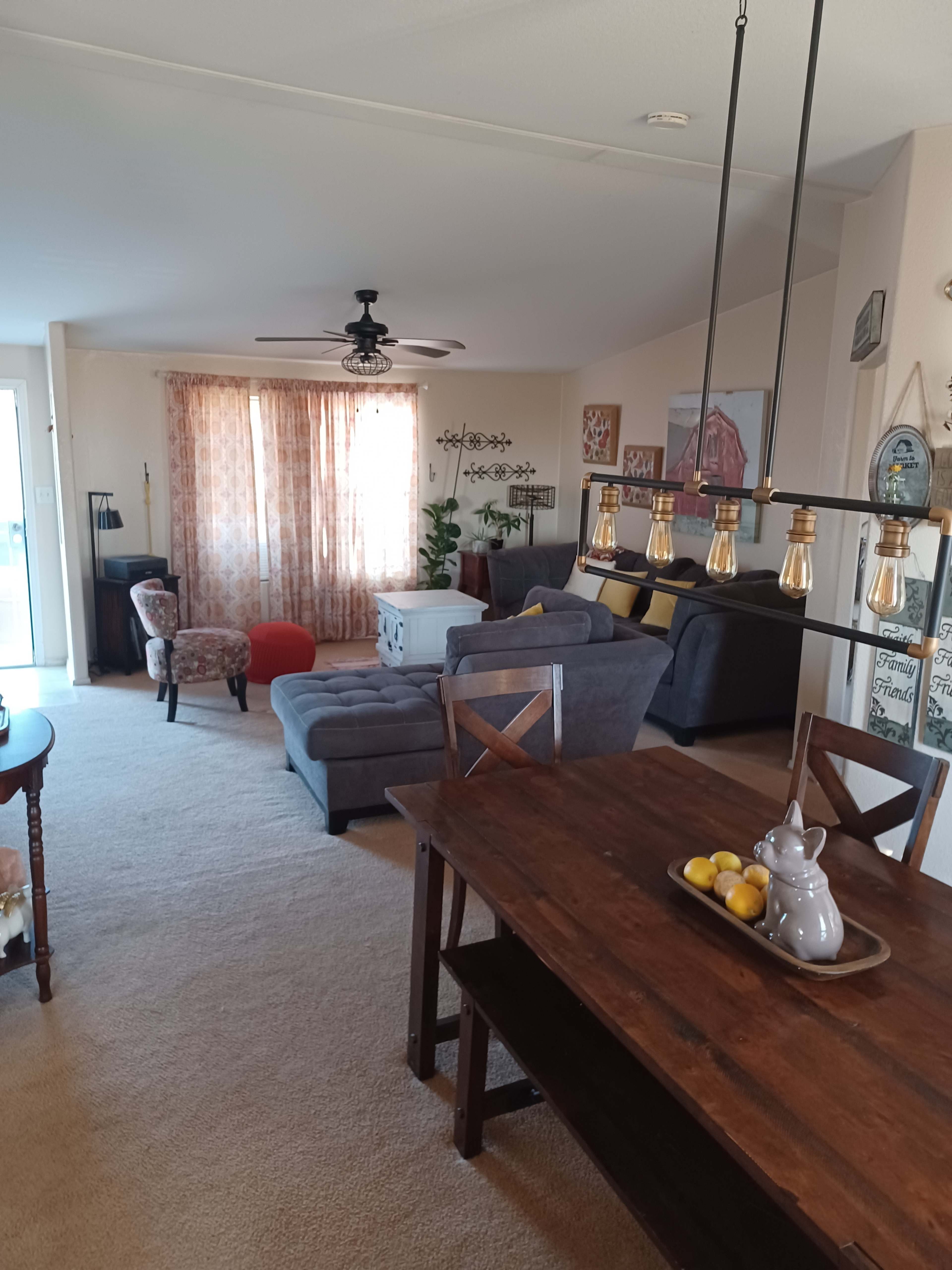 A cozy living room and dining area feature a combination of seating, a large window with curtains, and a wooden dining table with decorative items.