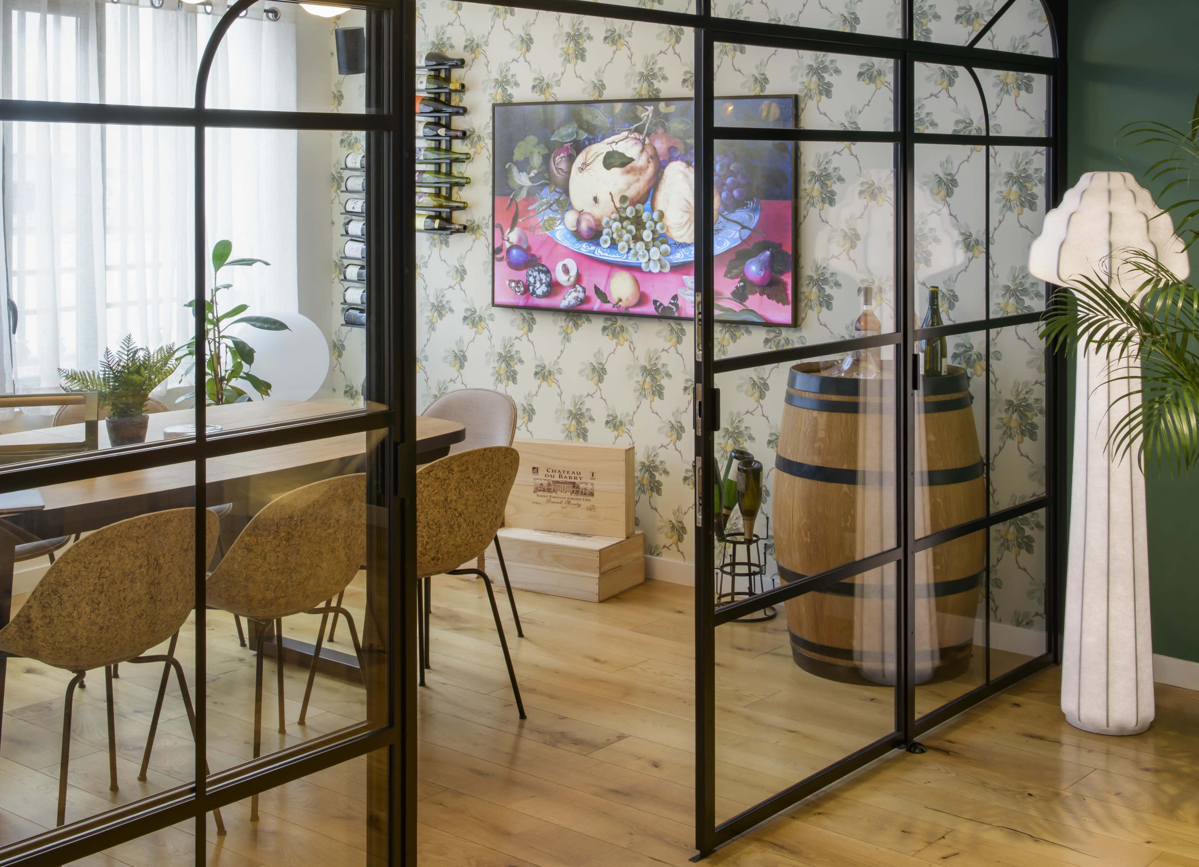 The image shows a stylish dining area separated by black-framed glass panels, featuring a wooden table, chairs, a barrel, and a wall adorned with floral wallpaper and artwork.