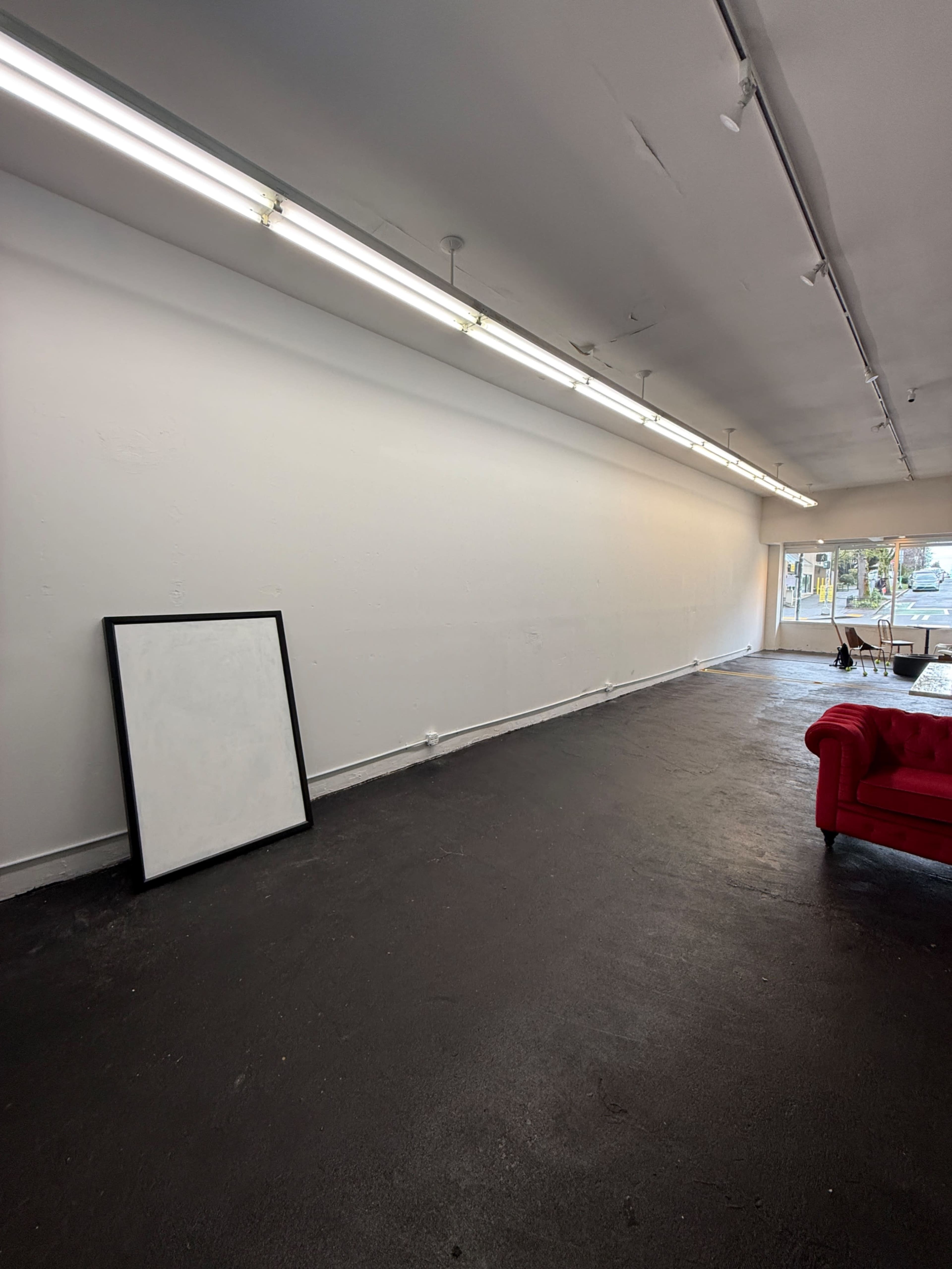 The image shows an empty interior space featuring a black floor, a large blank canvas resting against the wall, and a red sofa positioned nearby.
