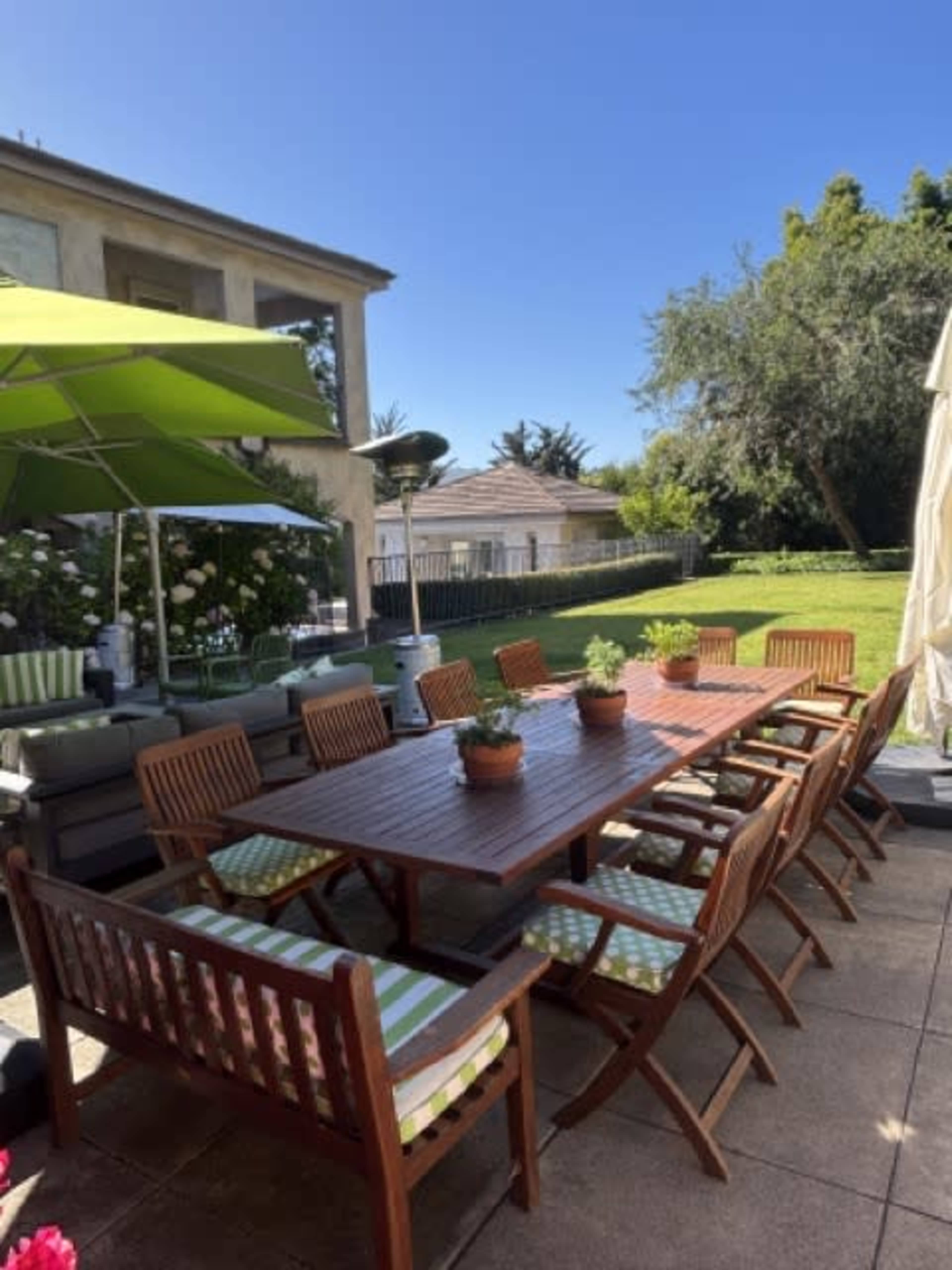A large wooden dining table surrounded by chairs is set up on a patio with a green umbrella, overlooking a grassy yard and nearby buildings.