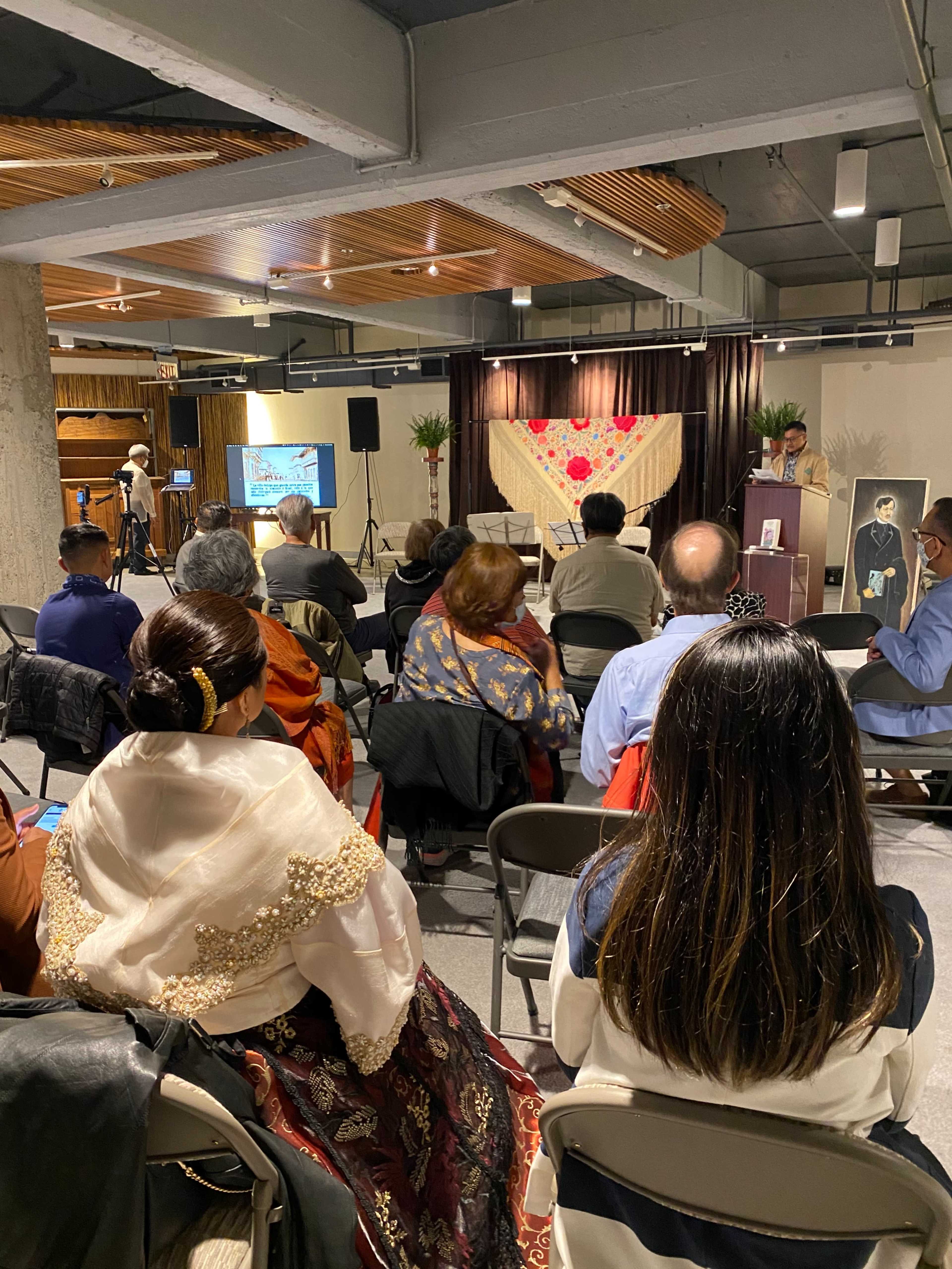 A diverse audience is seated in a spacious event room, where a speaker addresses them from a podium near a decorative backdrop.