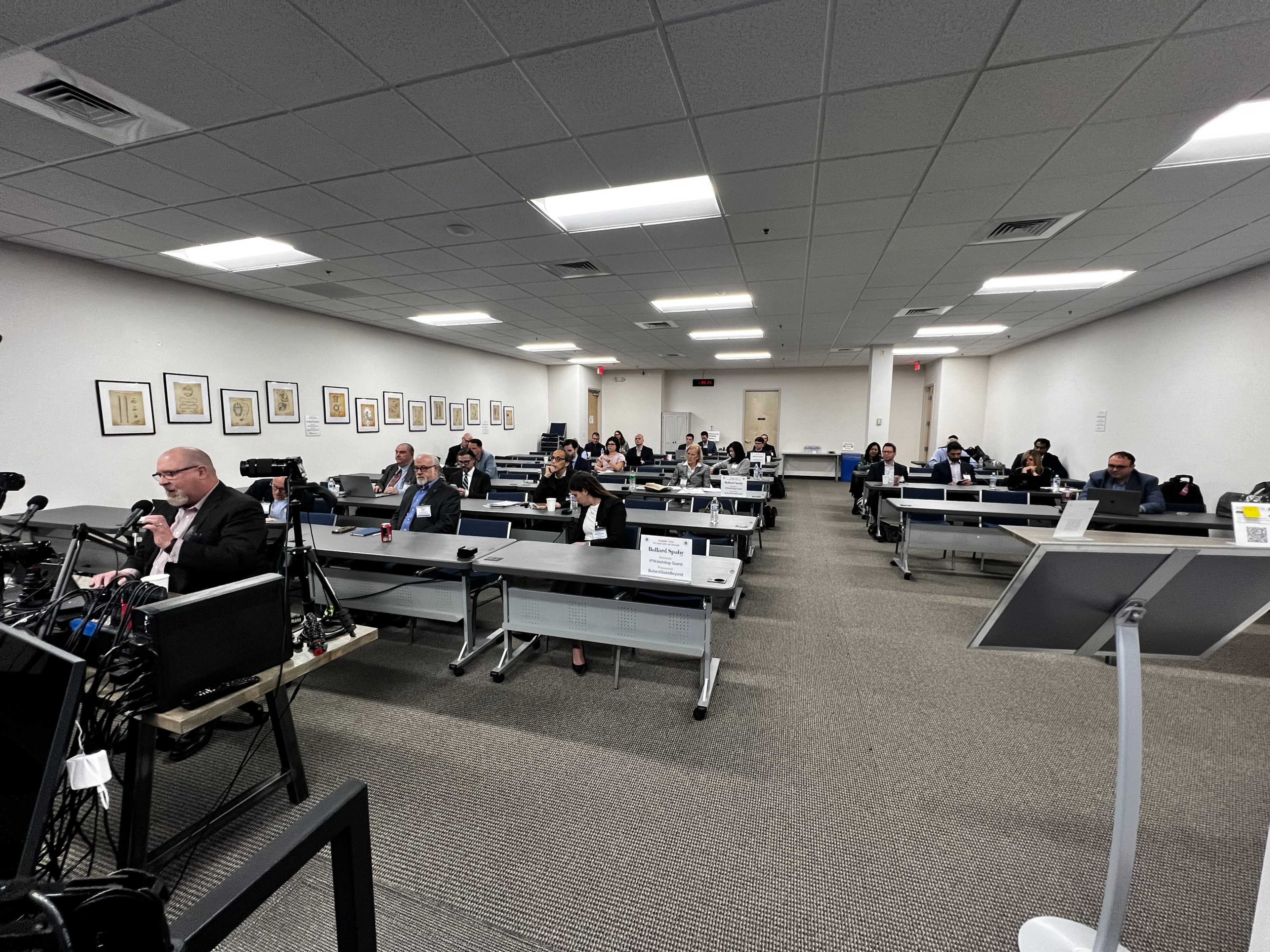 A large conference room is filled with seated participants attentively facing a speaker at the front, with microphones and equipment set up for a presentation.