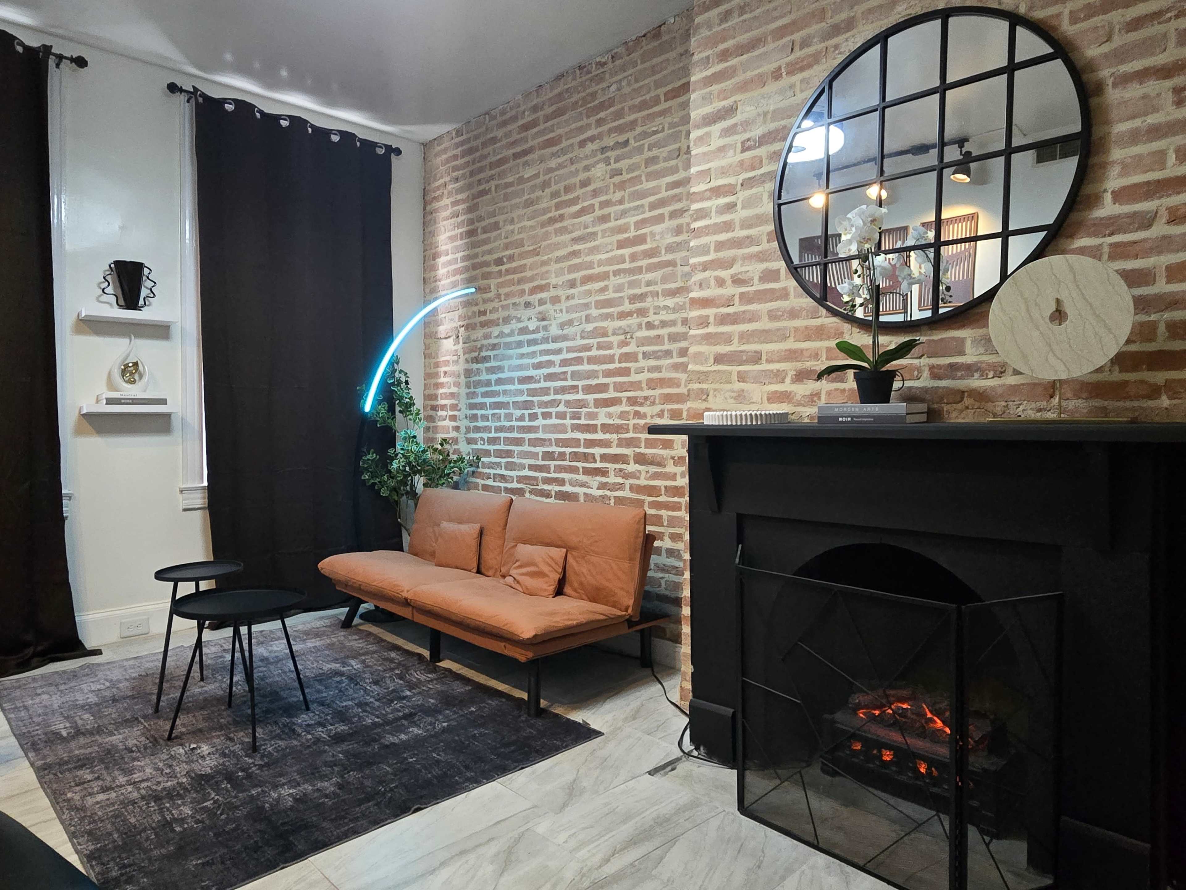 The image shows a cozy living room featuring a brick wall, a brown leather sofa, a small round table, and a fireplace with an electric fire effect.