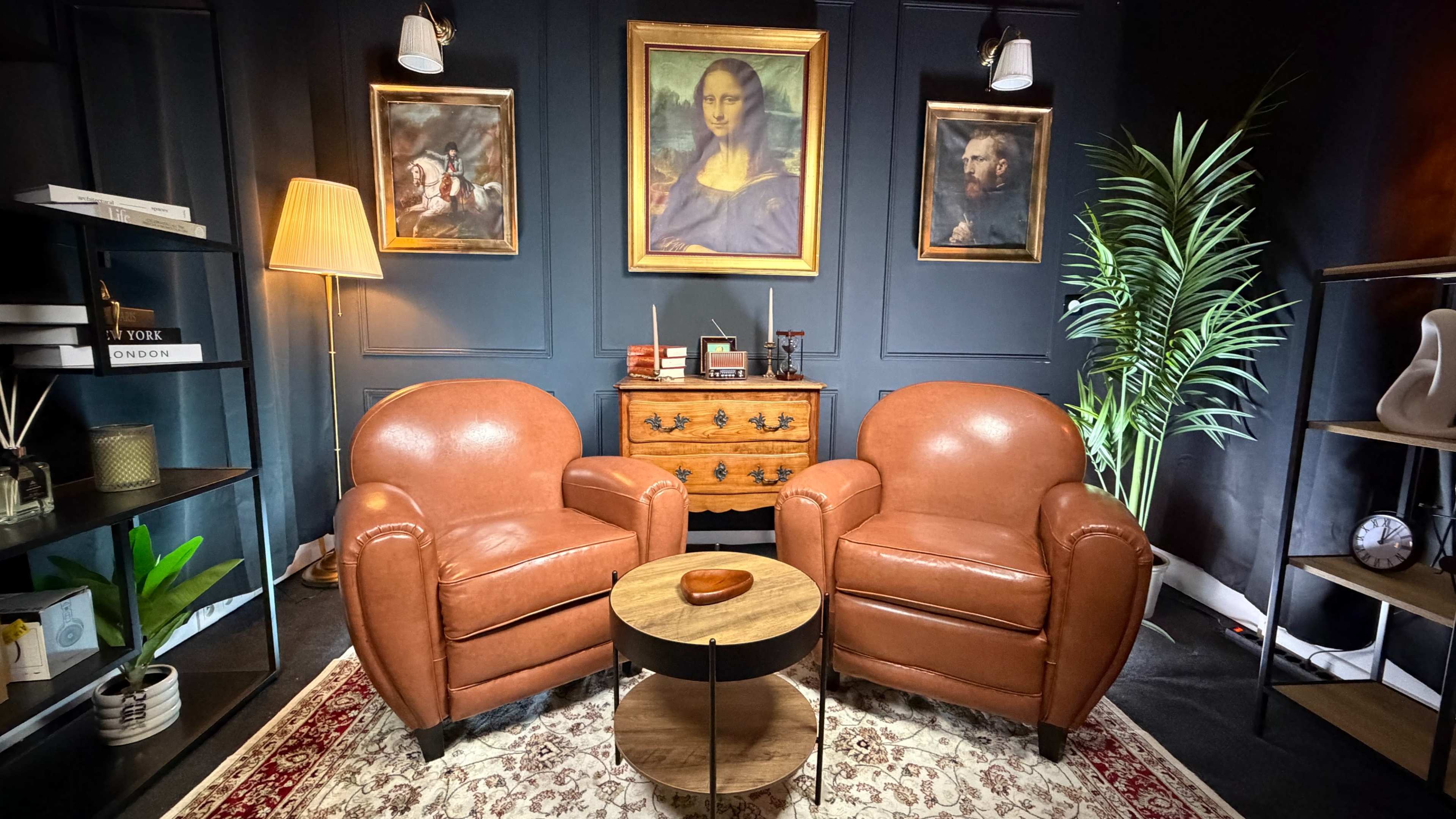 The image features two brown leather armchairs facing each other, with a round wooden coffee table in between them, set against a backdrop of dark blue walls adorned with paintings.