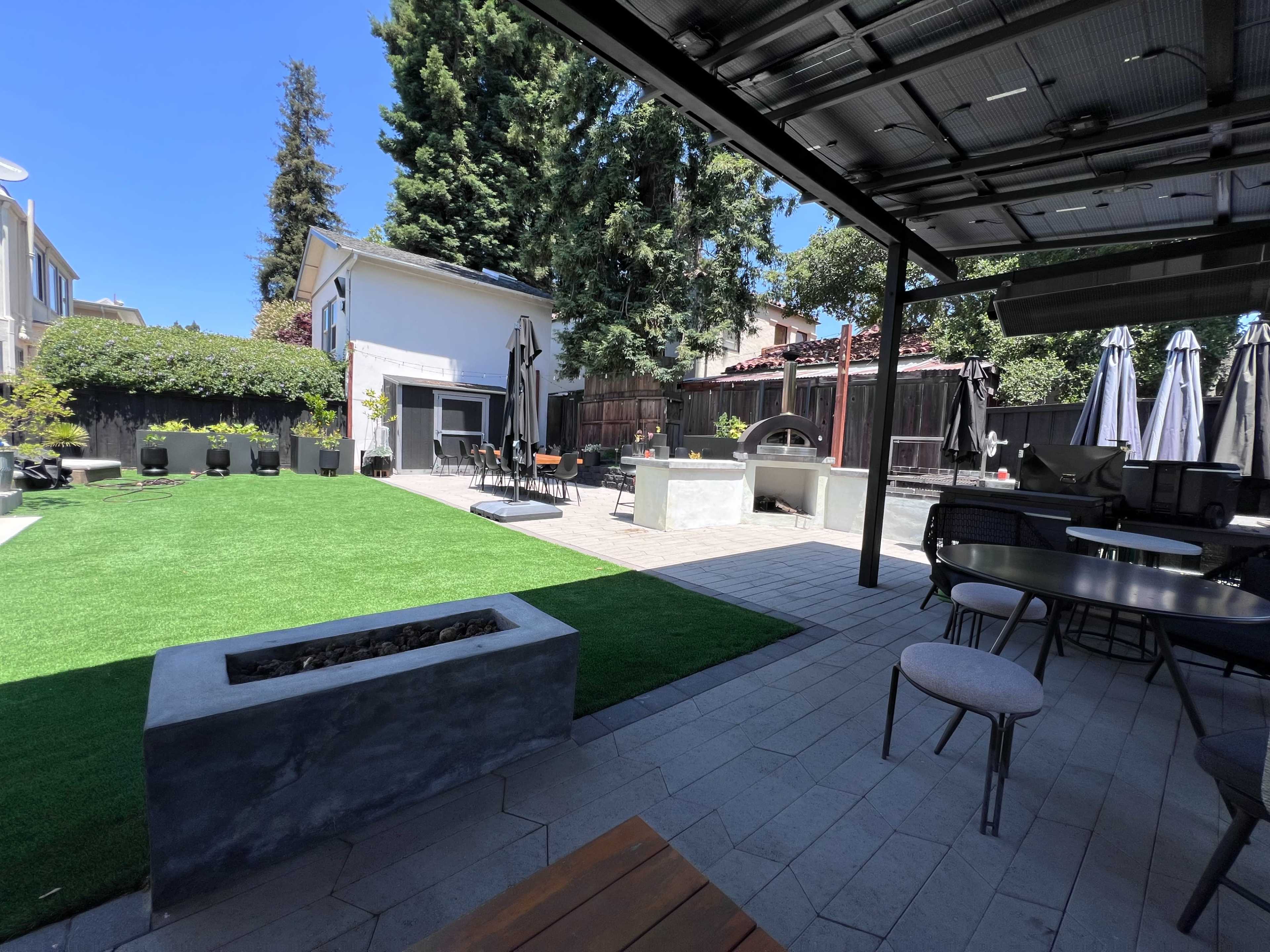 The image depicts a modern outdoor patio area featuring landscaped grass, seating arrangements, and a fire pit surrounded by tall trees and an adjacent building.