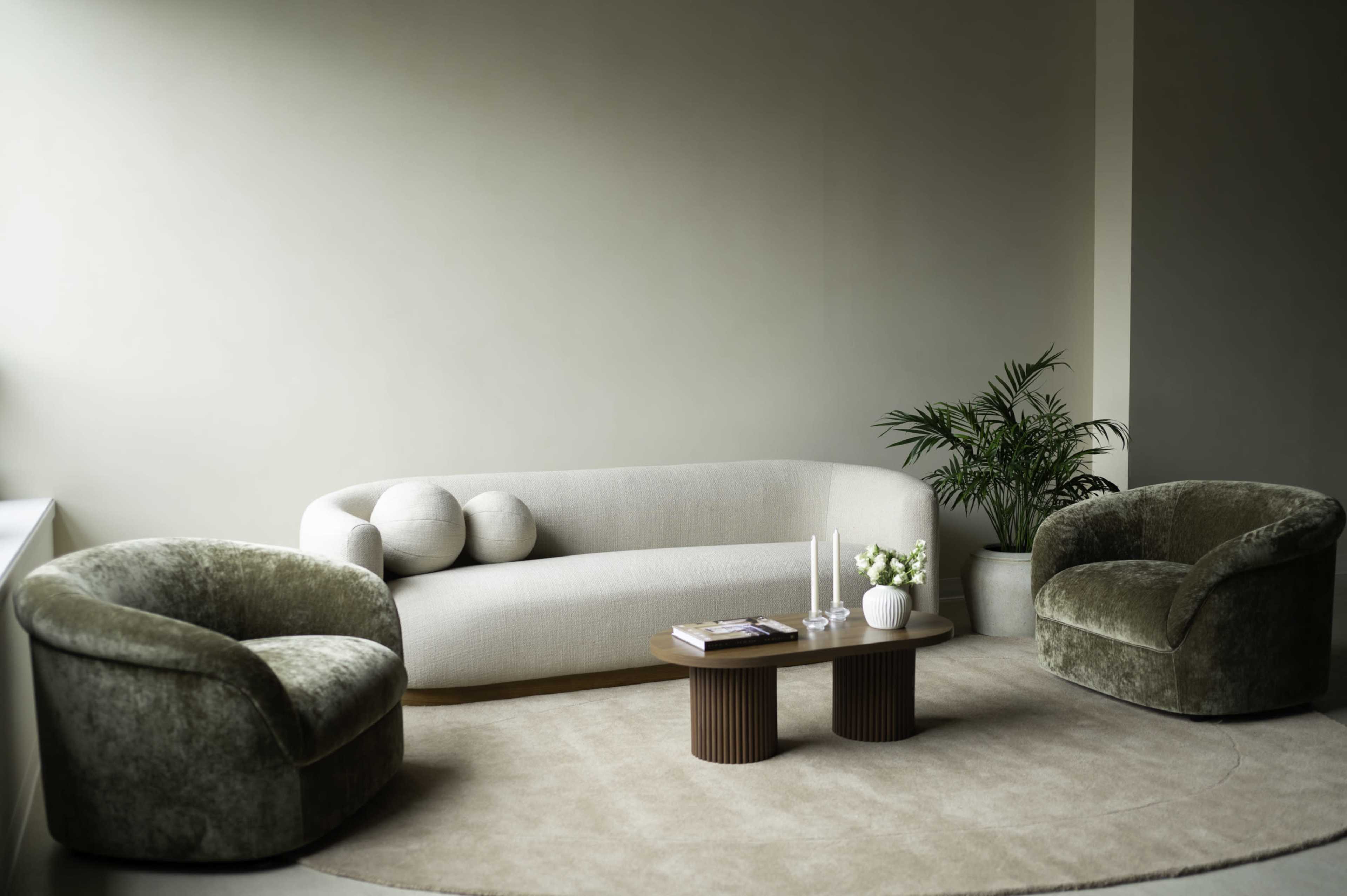 A modern living room features a light-colored sofa, two plush armchairs, a round coffee table, and a potted plant.