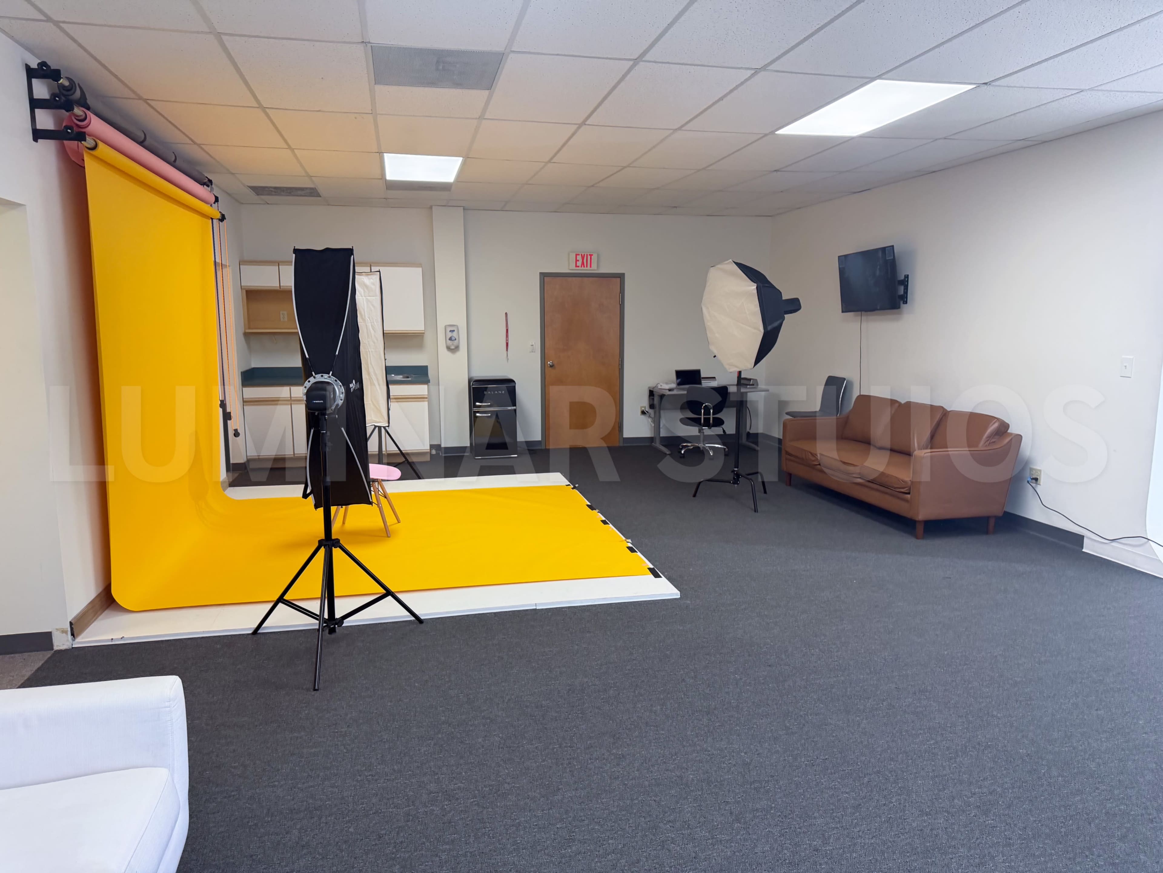 The image shows a photography studio with a bright yellow backdrop, lighting equipment, and a sofa arranged in a spacious room.
