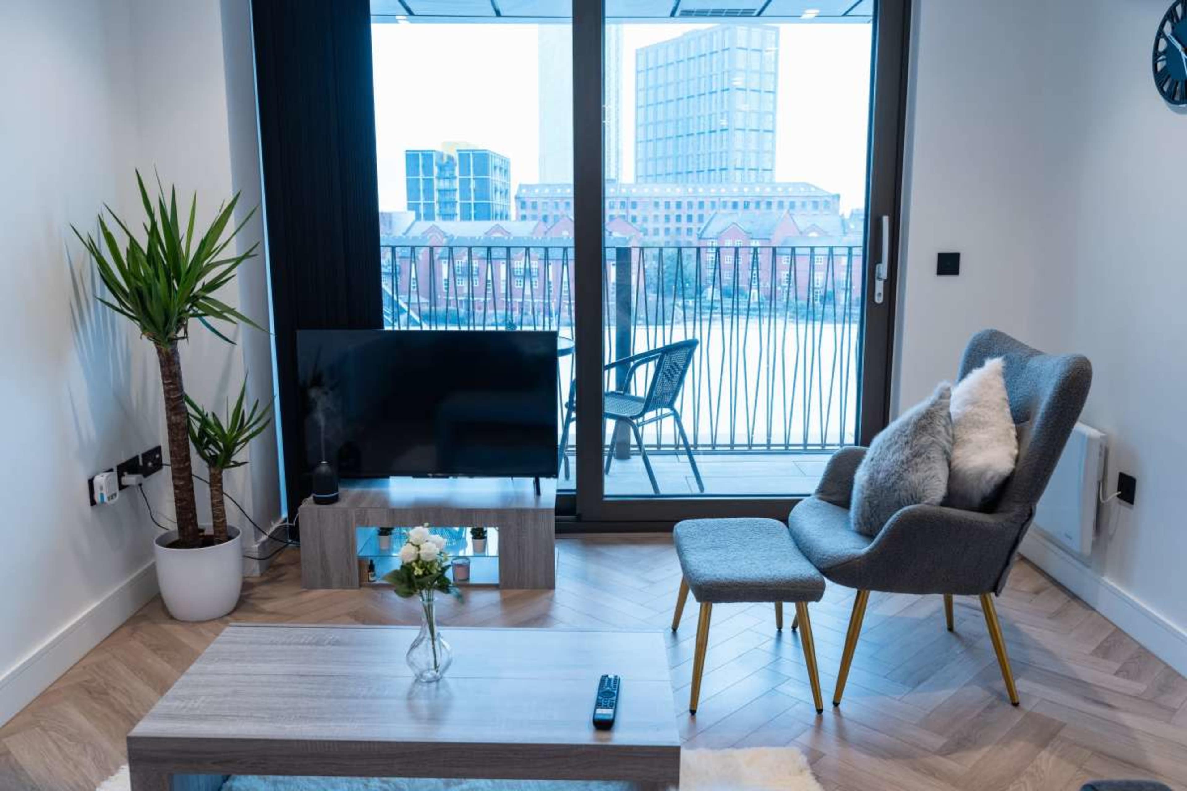 A modern living room features a chair, a table with a flower vase, a television, and a view of the outside through large glass doors.