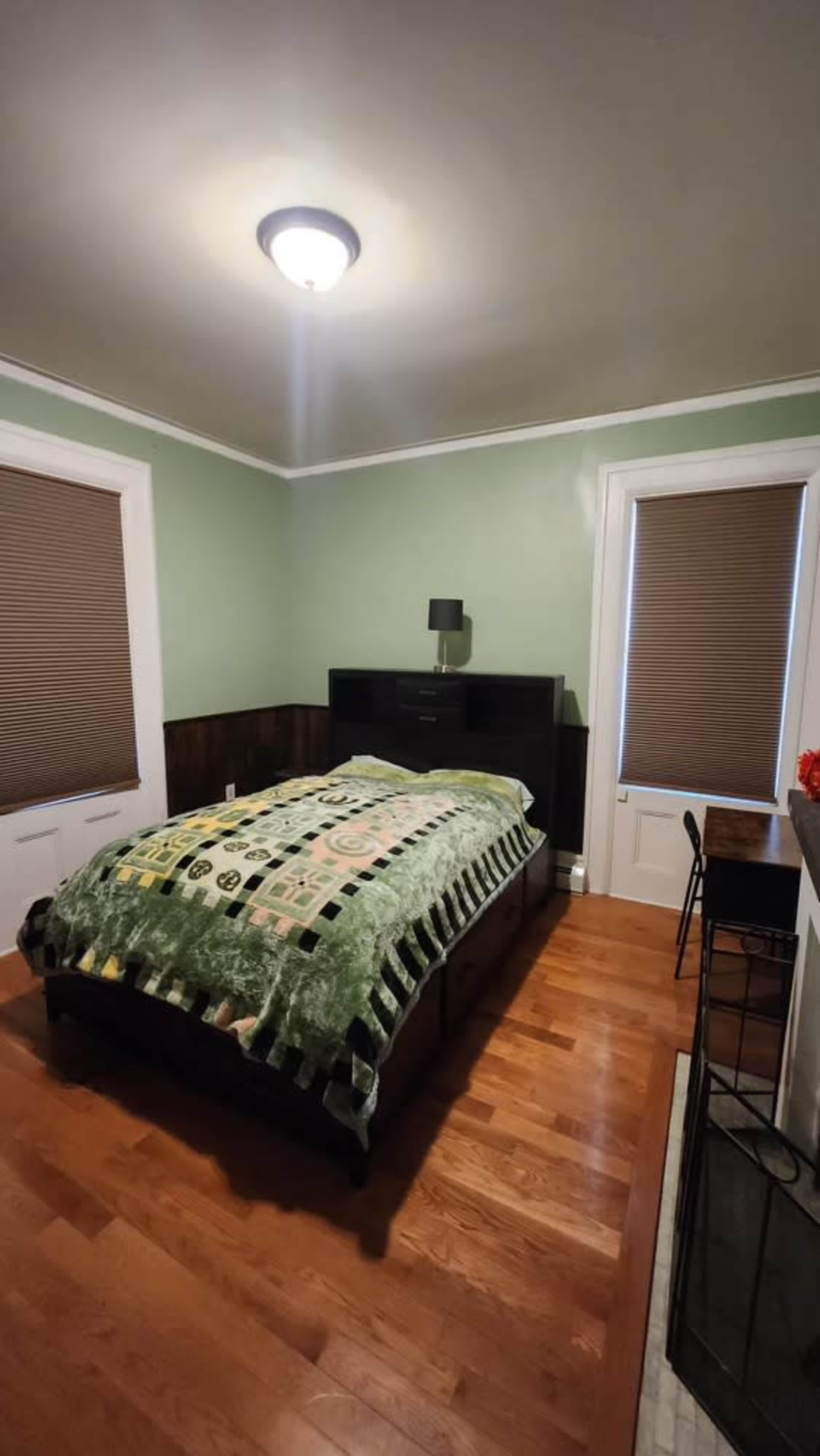 A neatly arranged bedroom features a bed covered with a green blanket, dark wood furniture, and two windows with blinds.