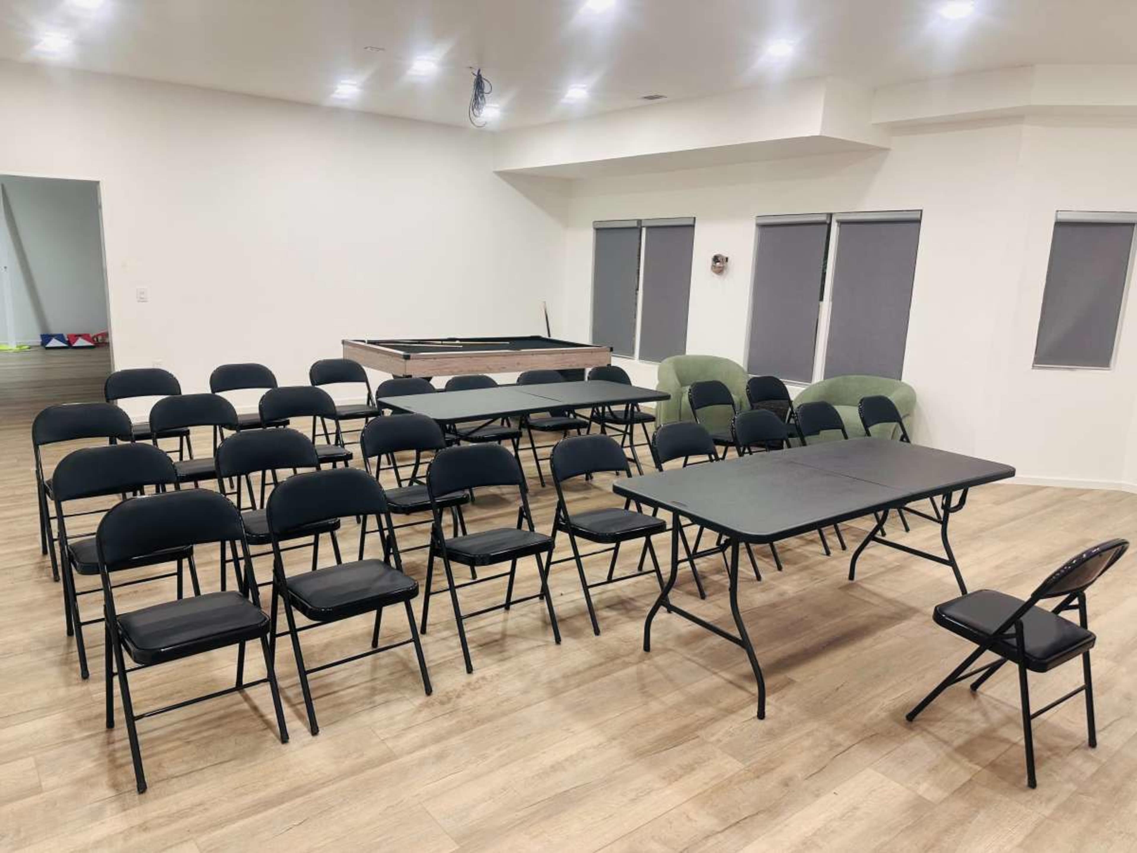 The image shows a spacious, well-lit room arranged with rows of black folding chairs and several tables, with some green chairs visible in the background.