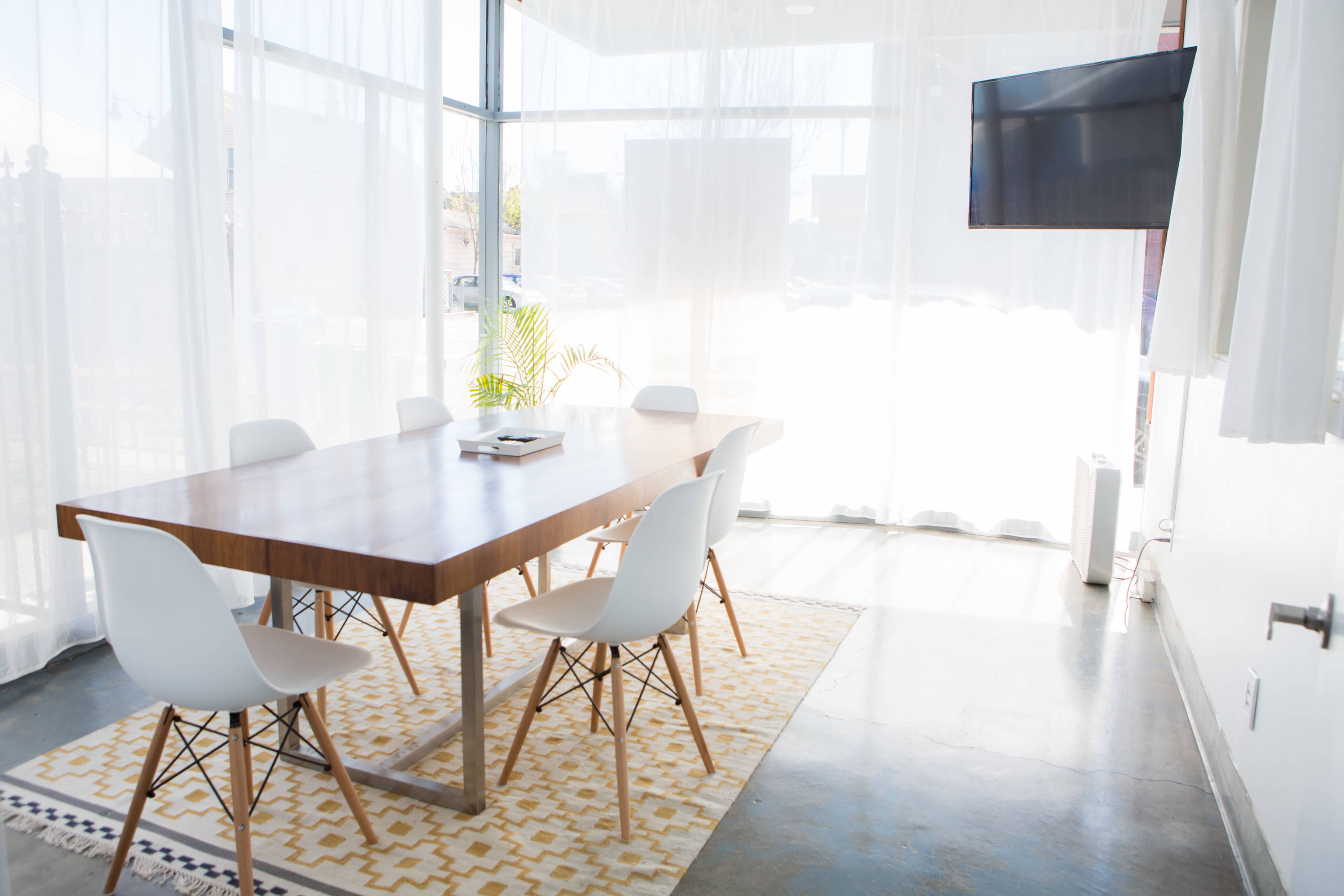 A modern conference room features a wooden table surrounded by white chairs, with large windows partially covered by sheer curtains.