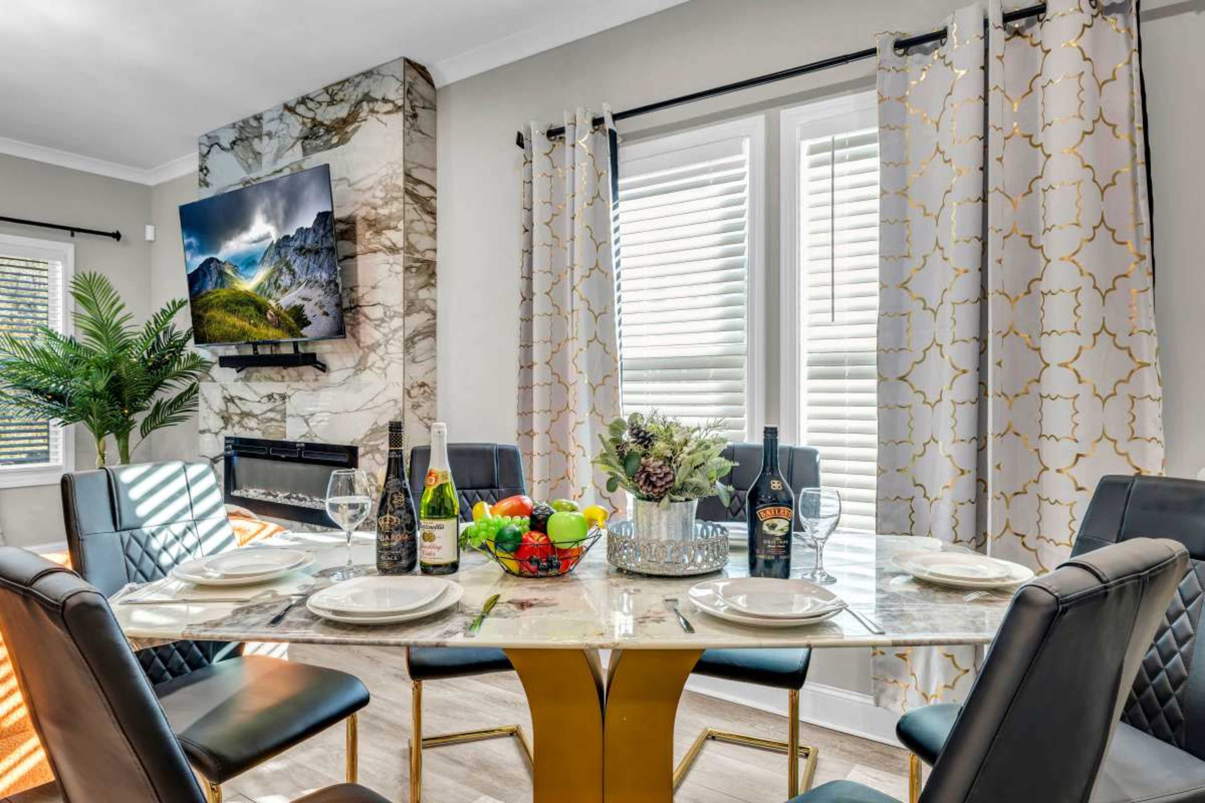 A dining table is elegantly set with plates, glasses, and fruit, surrounded by black chairs, under bright light filtering through sheer curtains beside a television mounted on a marble wall.