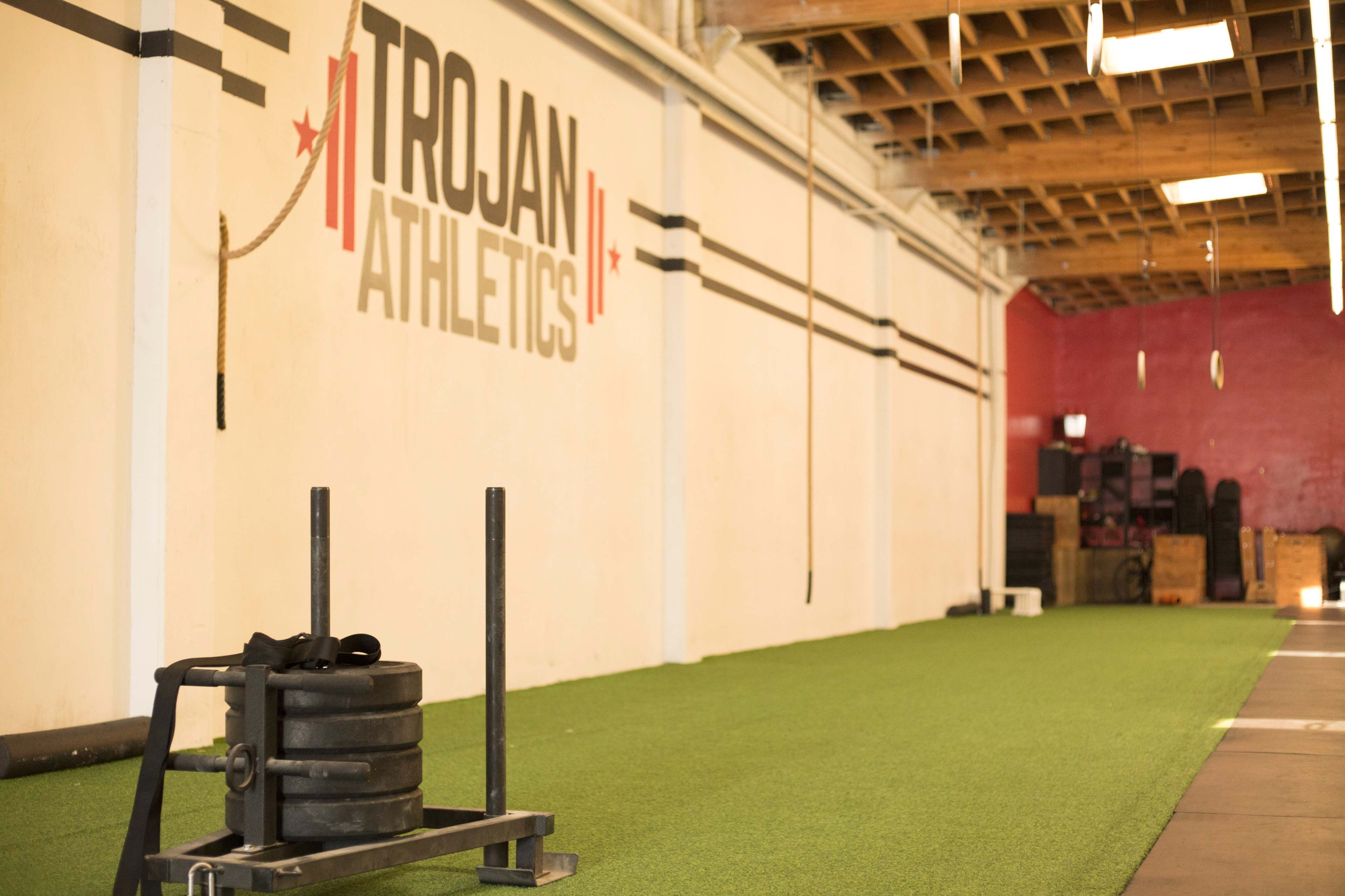 The image shows an indoor athletic training area with a green turf surface, a sled for weight training, and a wall featuring the logo "TROJAN ATHLETICS."