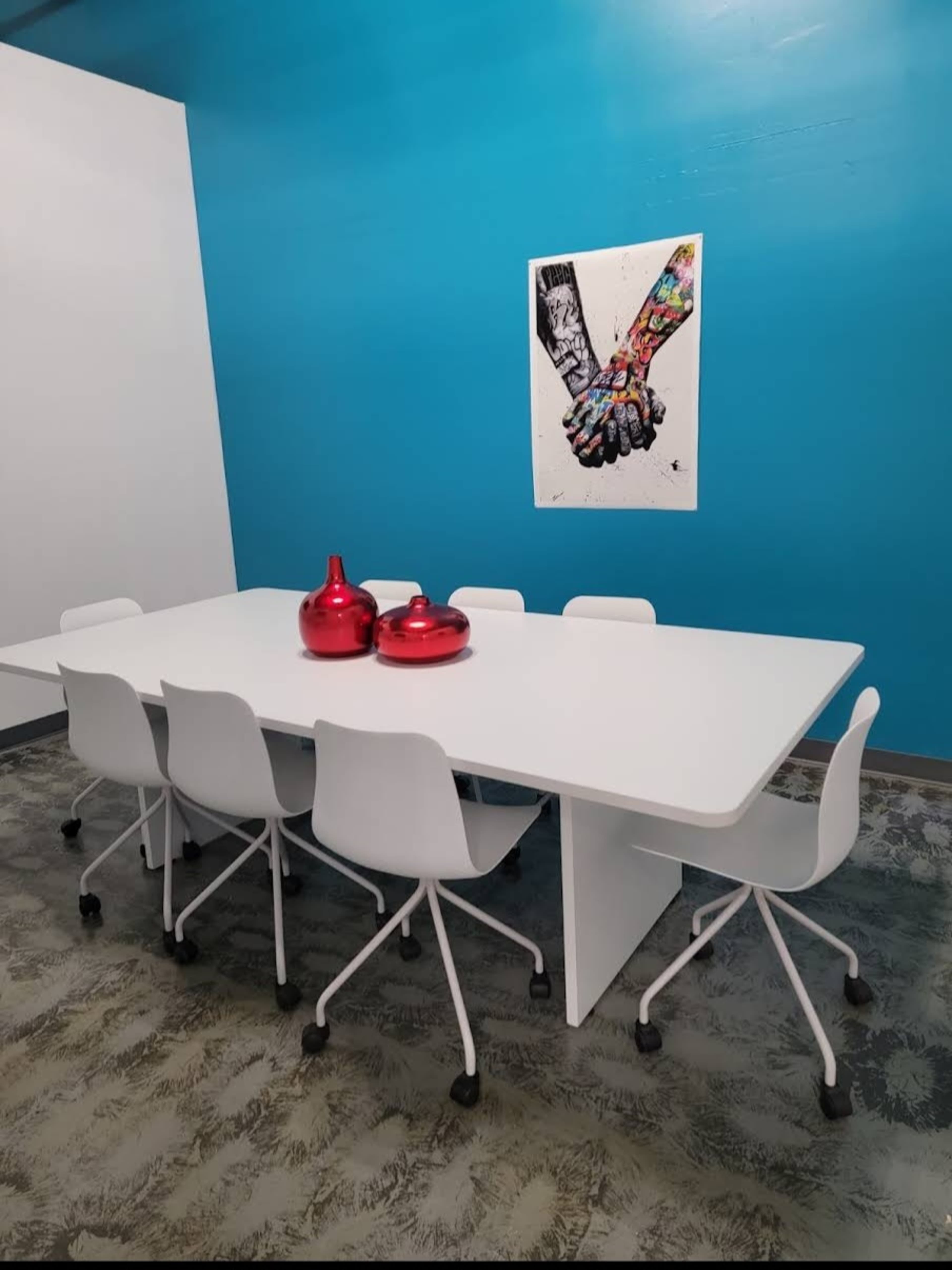 A modern conference room features a large white table surrounded by white chairs, with two red decorative vases and a colorful artwork of hands on the blue wall.
