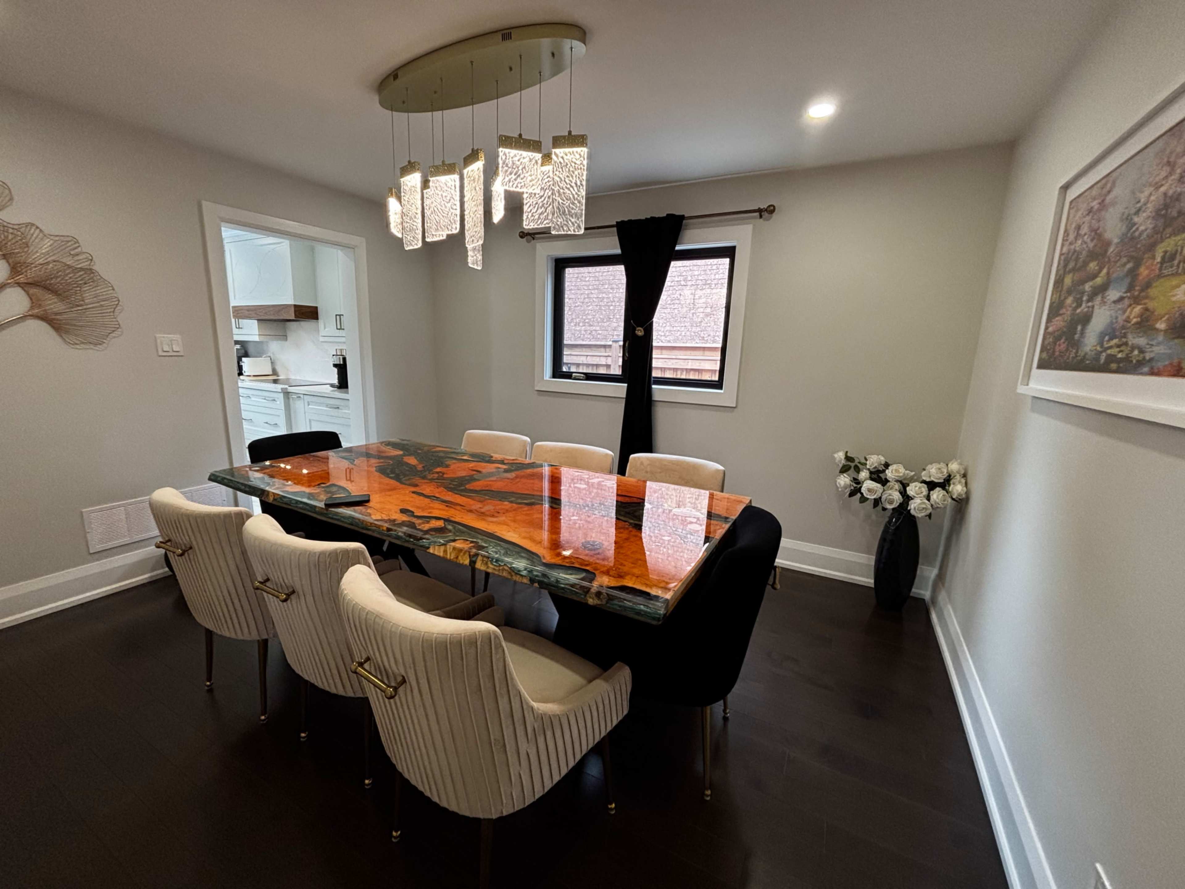 The image shows a dining room featuring a large, orange and black table surrounded by six upholstered chairs, with a modern light fixture above and a window allowing natural light in.
