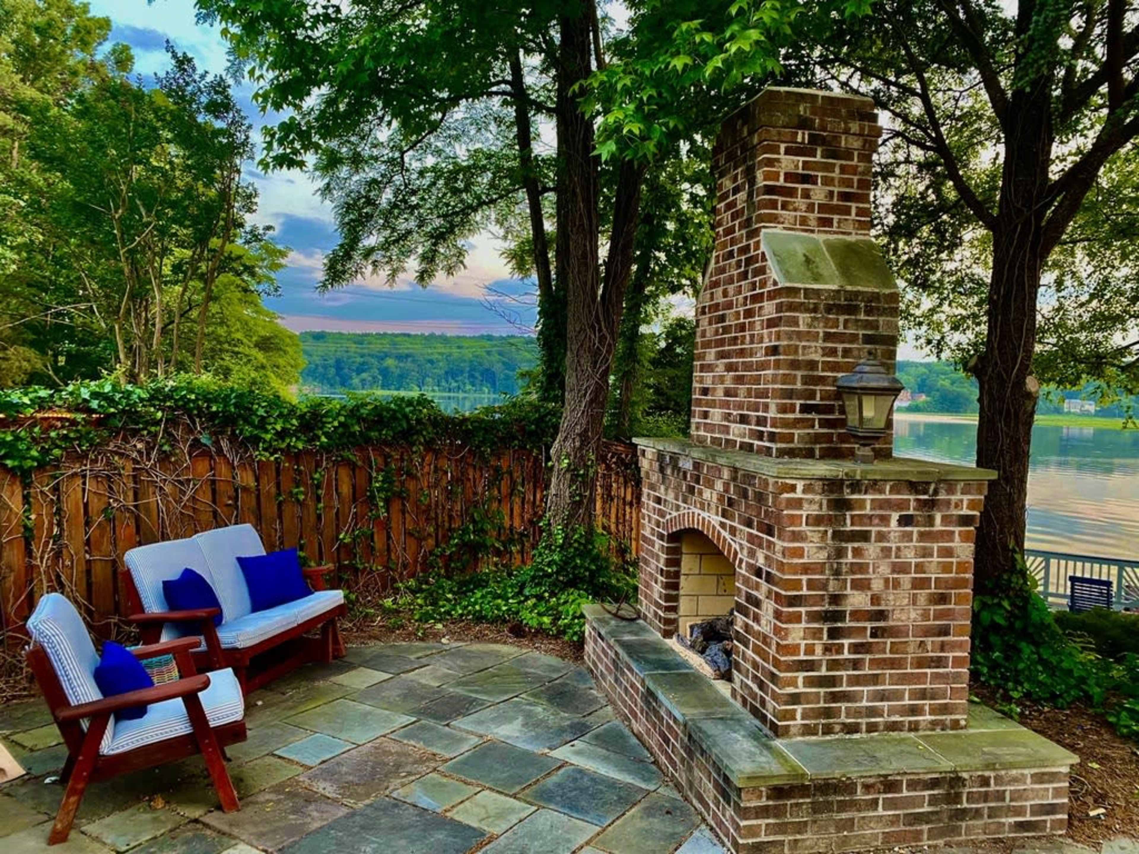 A stone patio area with a brick outdoor fireplace, two chairs, and a backdrop of trees and a lake under a pastel sky.