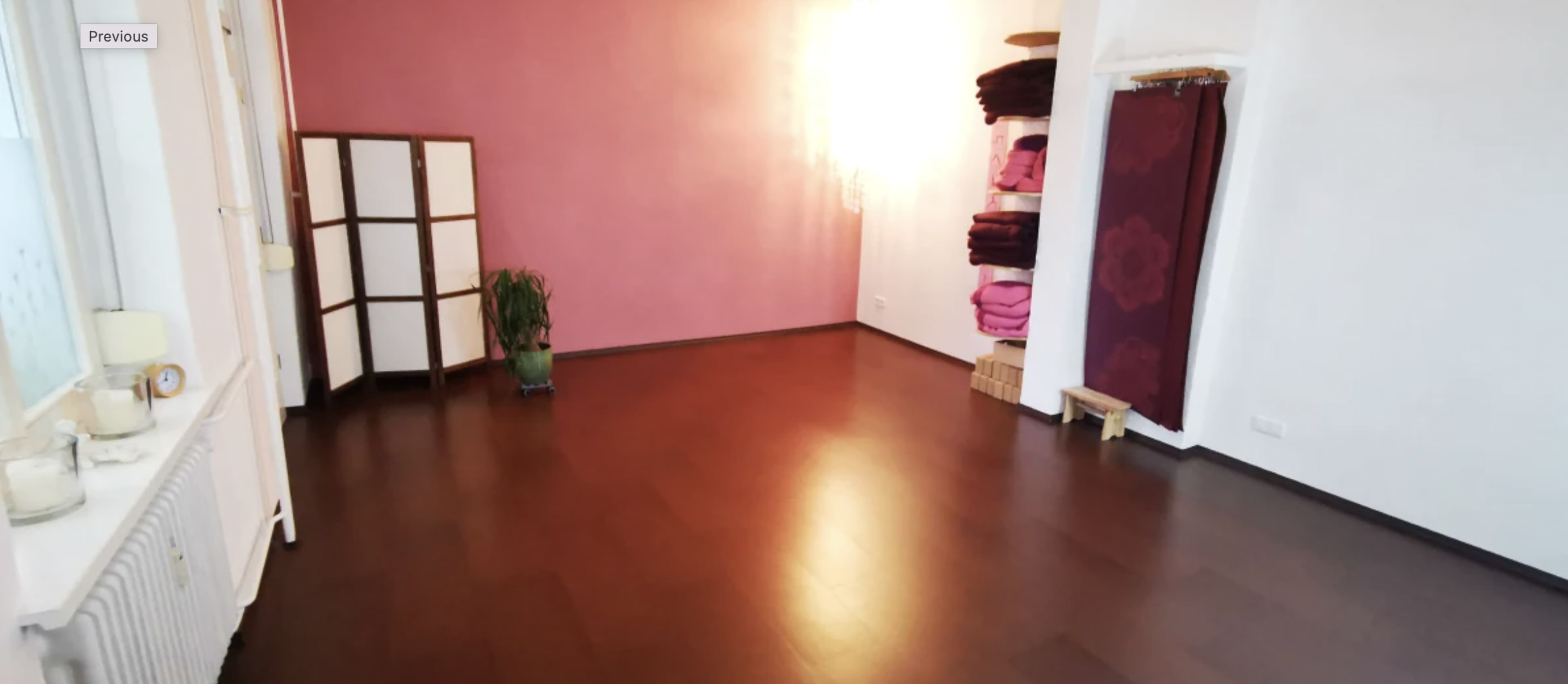 An empty room features a pink wall, a potted plant, a folding screen, and neatly stacked towels on a shelf.